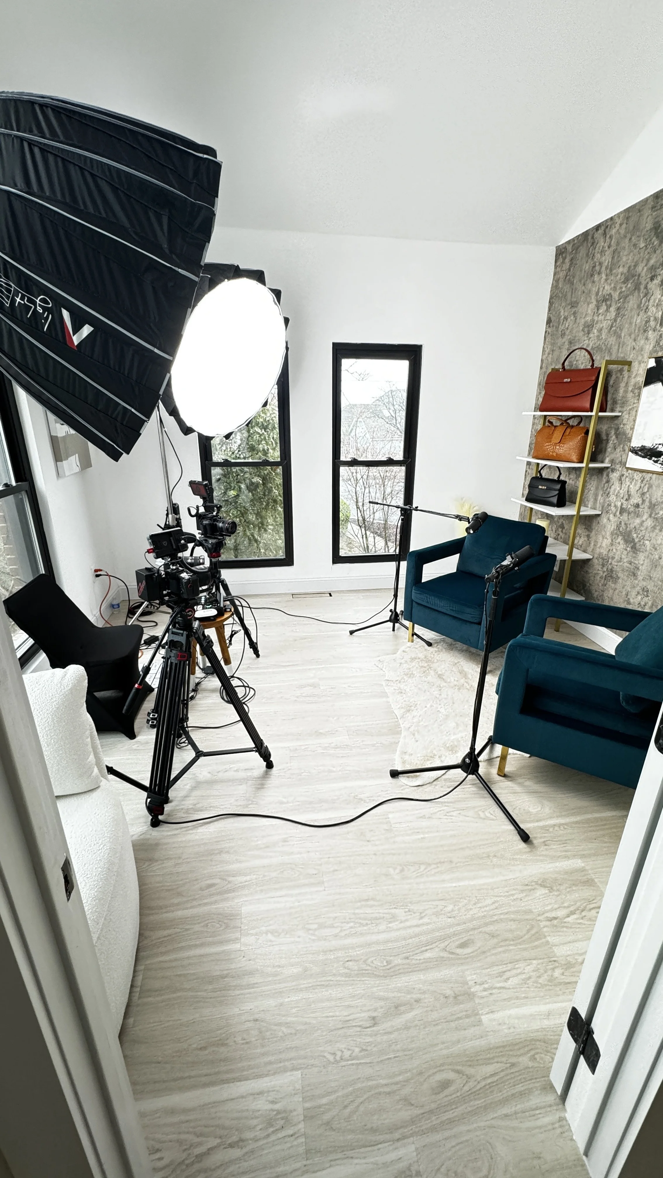 A small indoor filming setup with a camera on a tripod, two teal armchairs, a white sofa, and shelves with handbags. There are studio lights, microphones, and two large windows in the background.