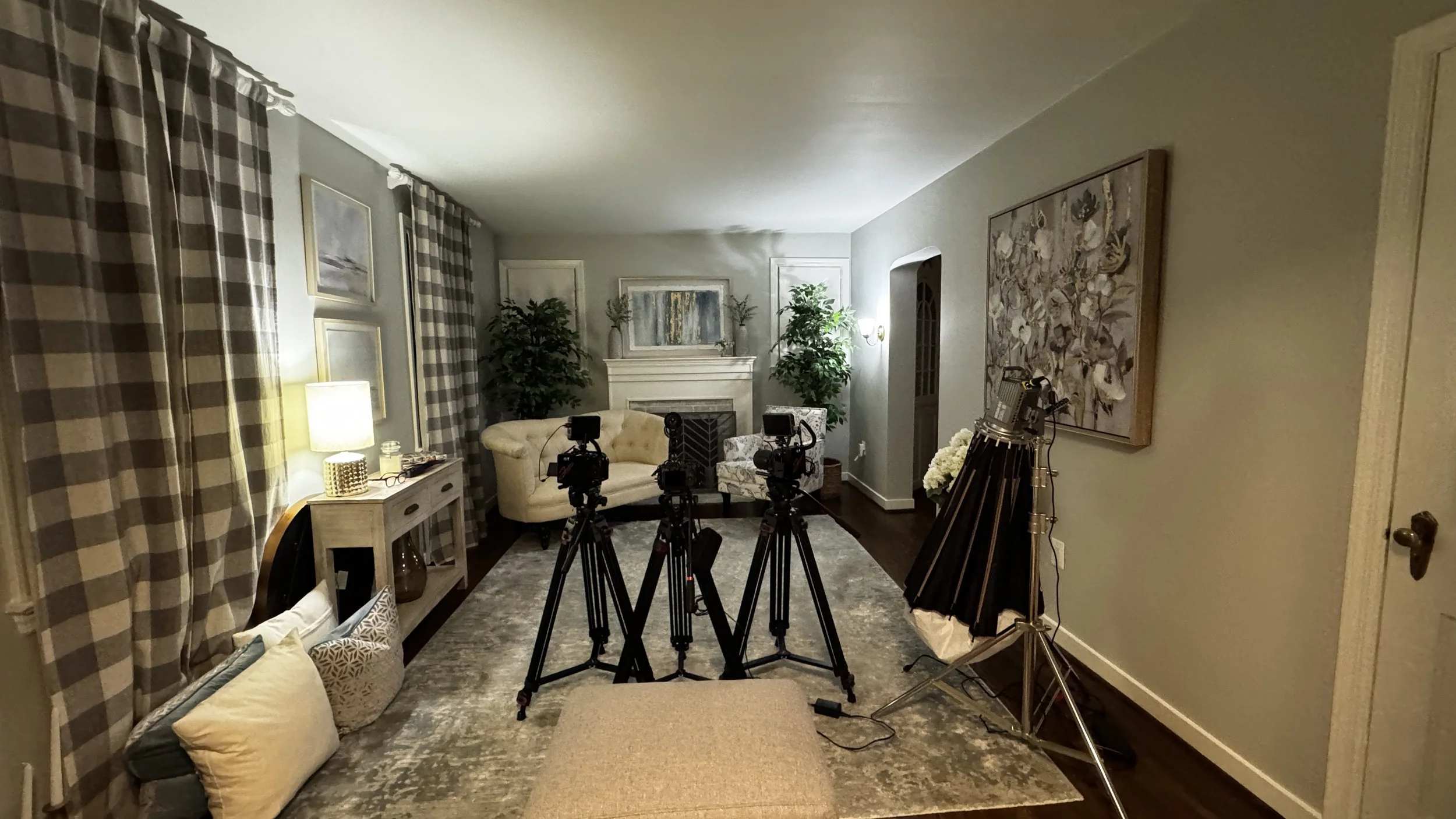 Living room set up for filming or photography, with three cameras on tripods facing the seating area, which includes a cream tufted sofa, patterned armchair, and side tables. There are framed wall art, checkered curtains, plants, a fireplace, a rug, 