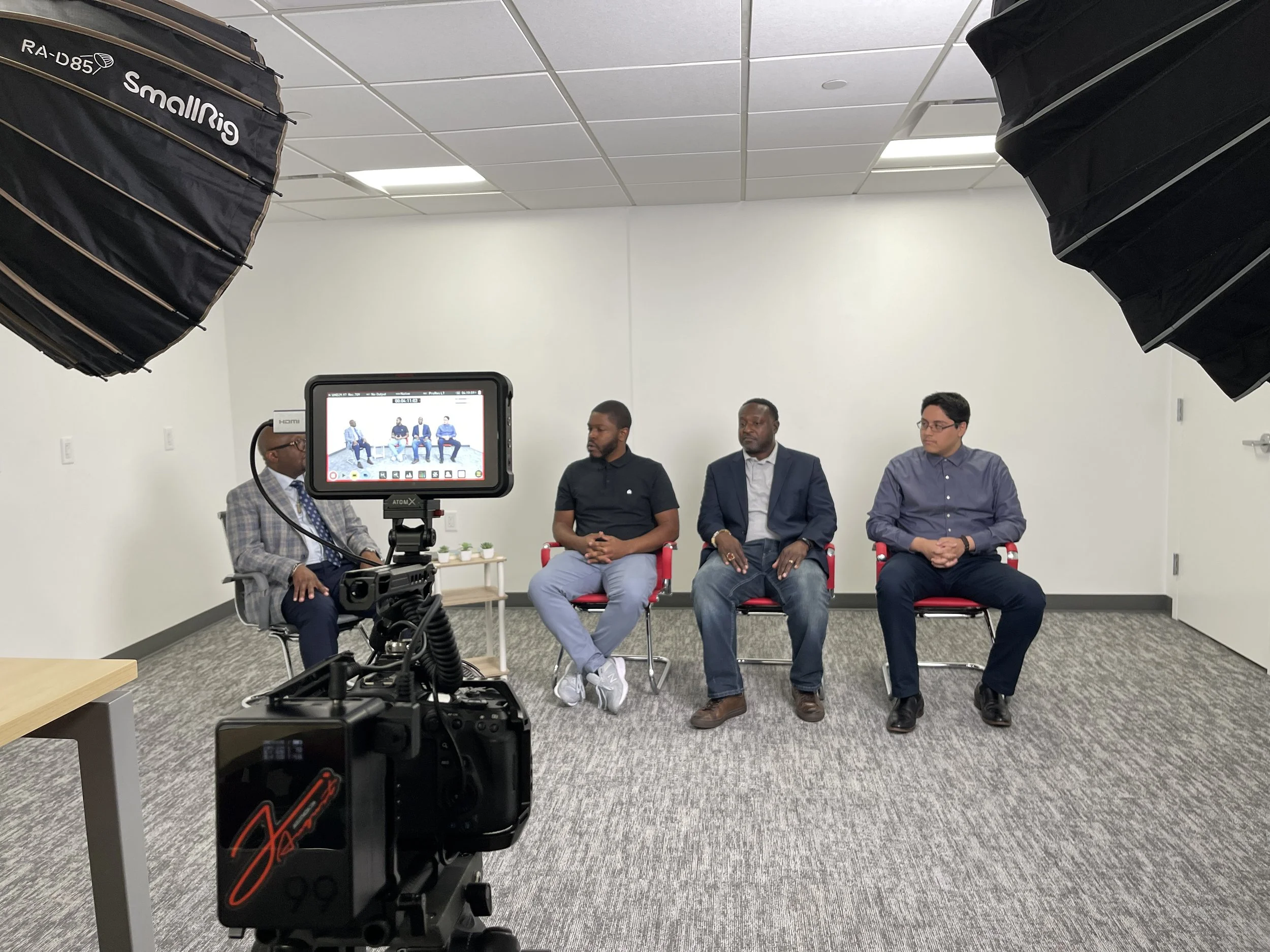 Four men sitting on chairs during a panel discussion or interview in a room with professional lighting and camera equipment.