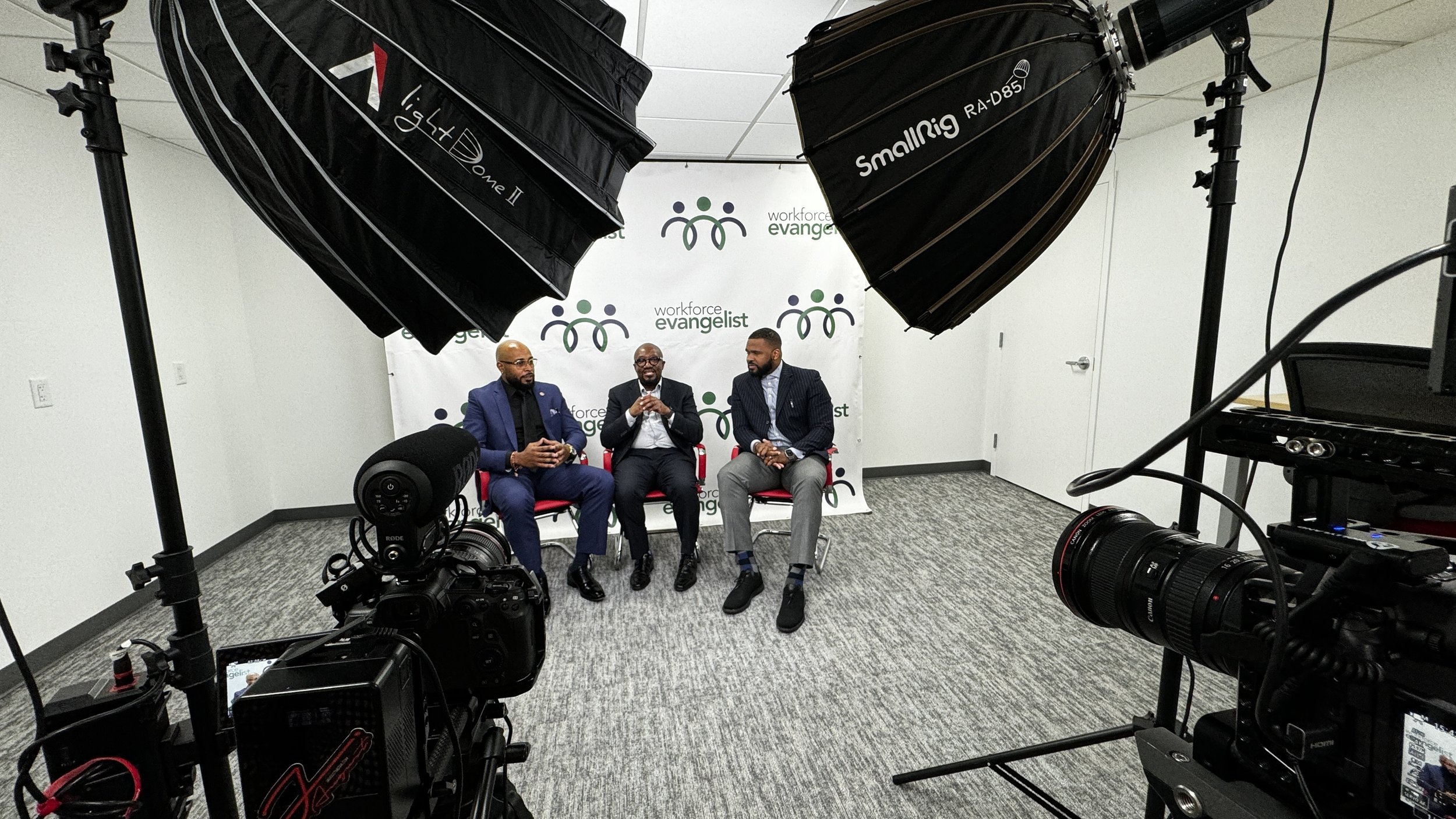 Three men seated on red chairs in a recording studio, engaged in a discussion or interview, with a backdrop branded 'Workforce Evangelist'; professional video cameras and lighting equipment surround them.