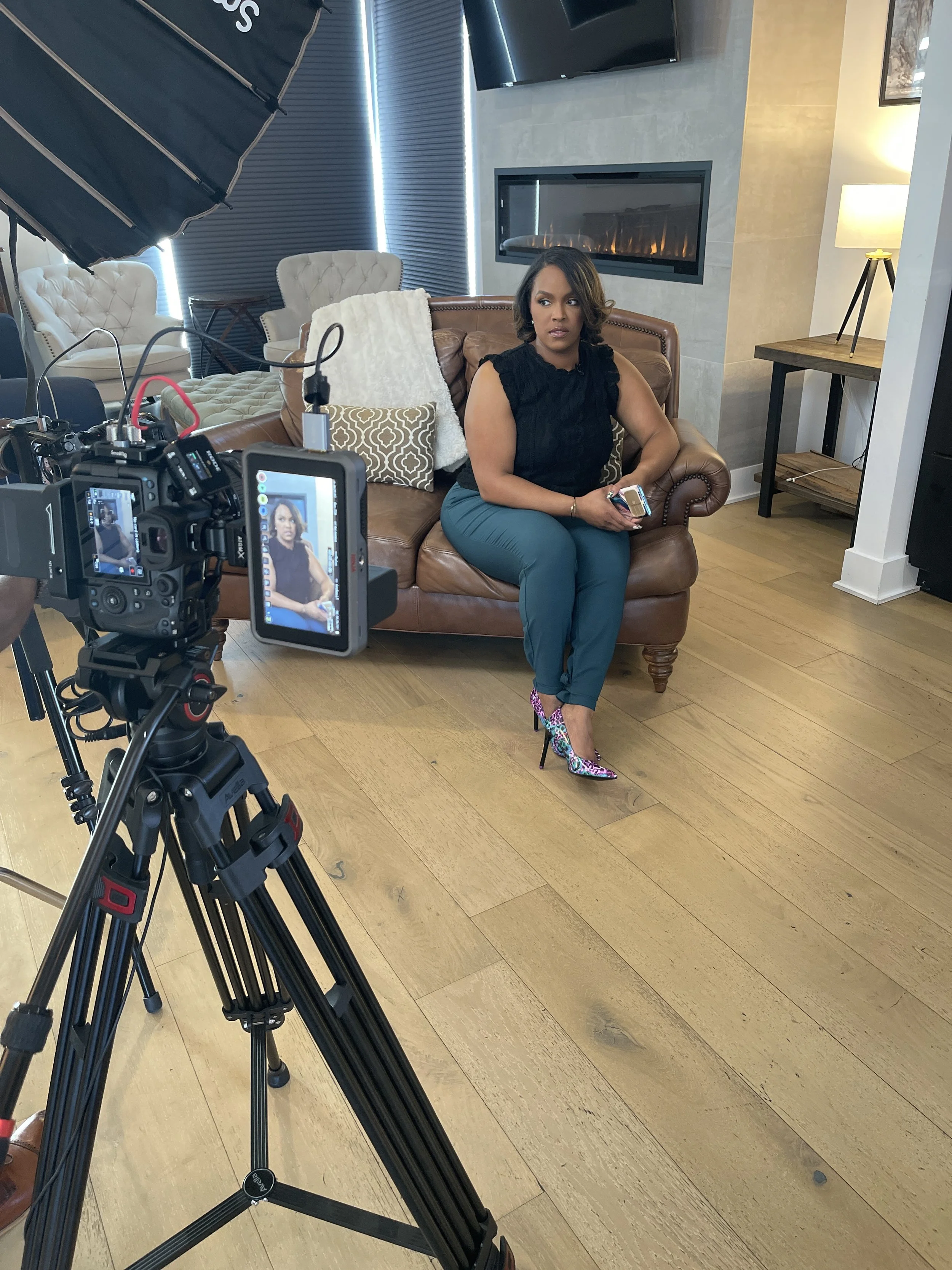 Woman sitting on a brown leather couch during a video shoot or interview, holding a phone, with camera and lighting equipment setup around her.