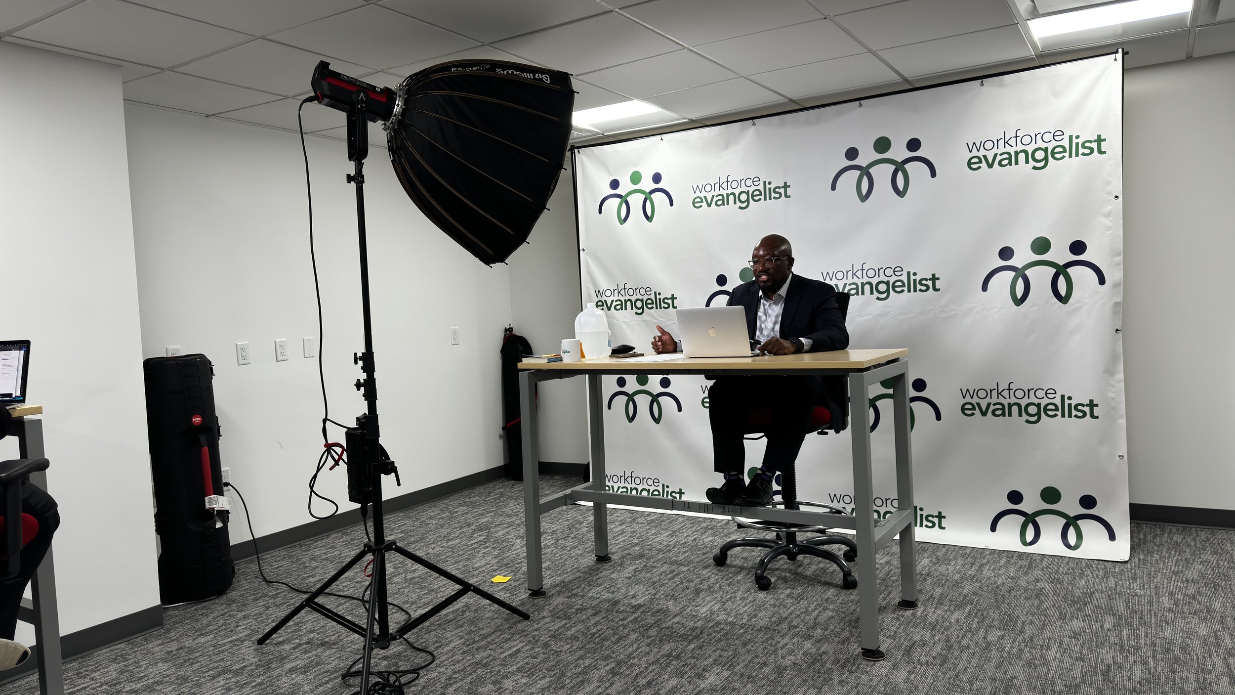 A man in a suit sitting at a desk with a laptop, in a studio with a backdrop reading 'workforce evangelist', and a large studio light to the left.