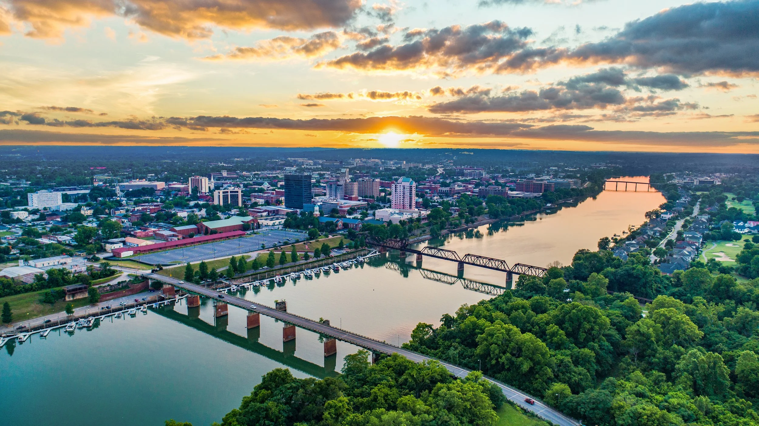 Aerial view of a city at sunset showing a river with a bridge, city buildings, and green trees along the riverbanks.