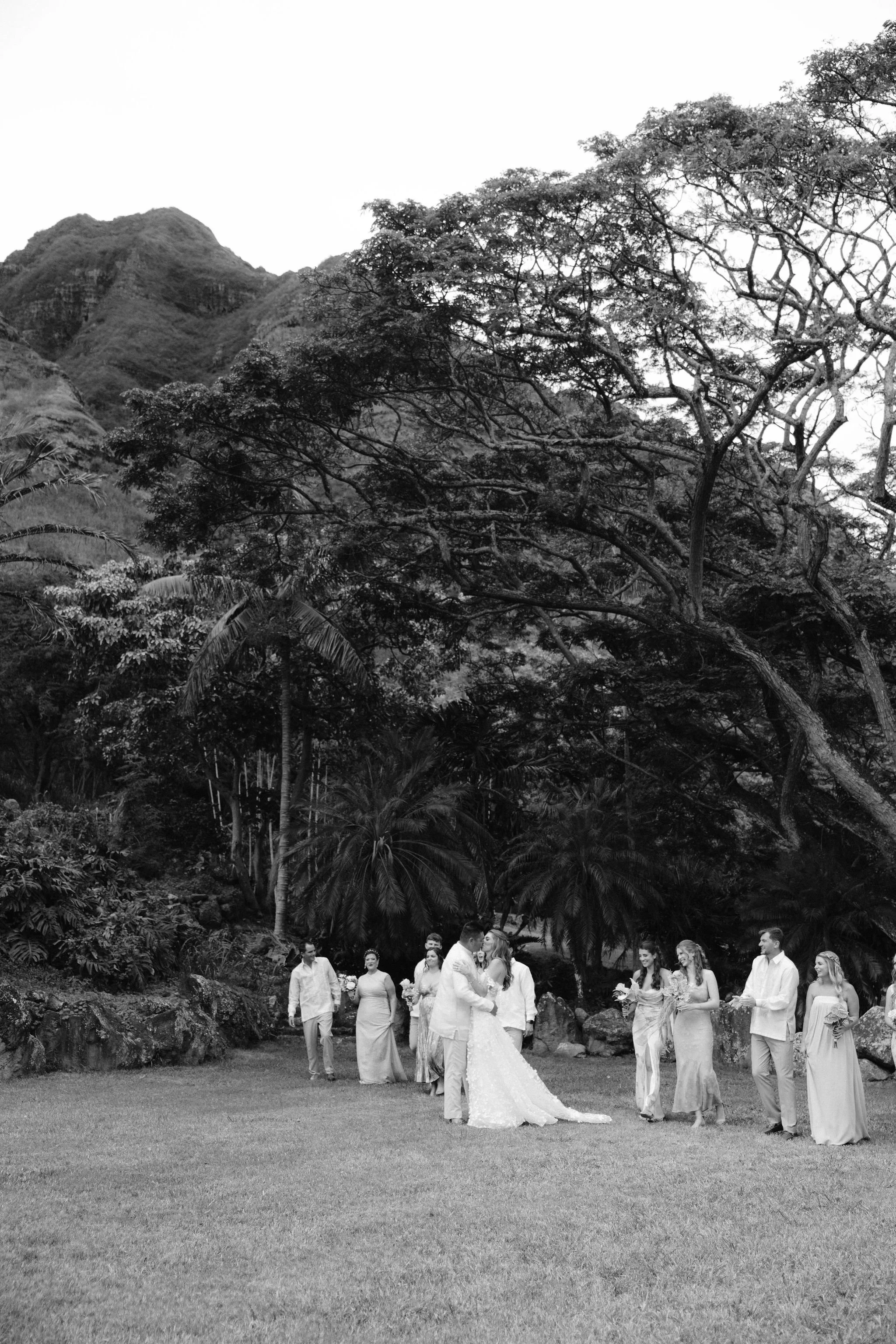 bride and groom kiss surrounded by their wedding party with kualoa ranch mountains in the background