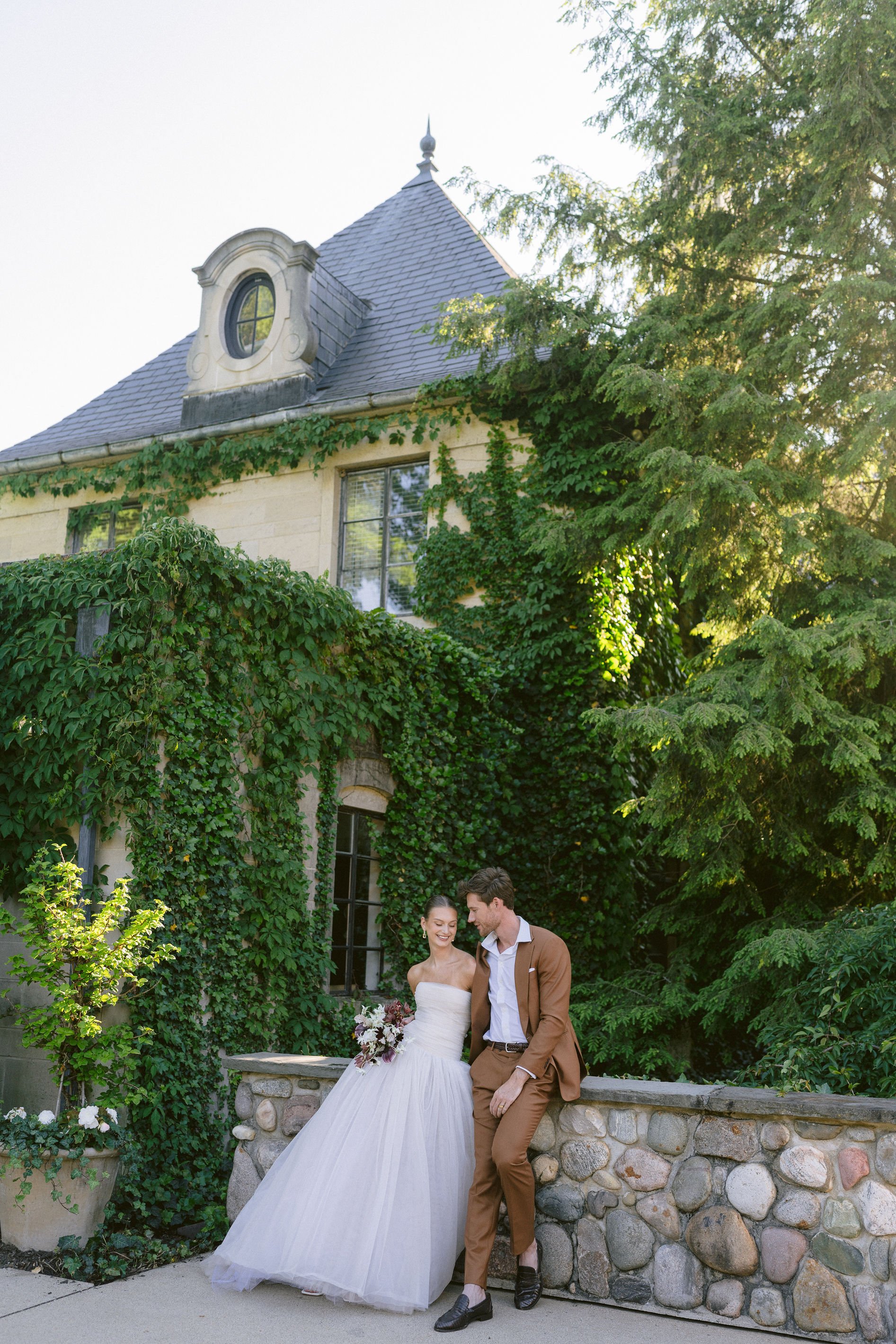 bride and groom lean against a rock ledge with greencrest manor in the background
