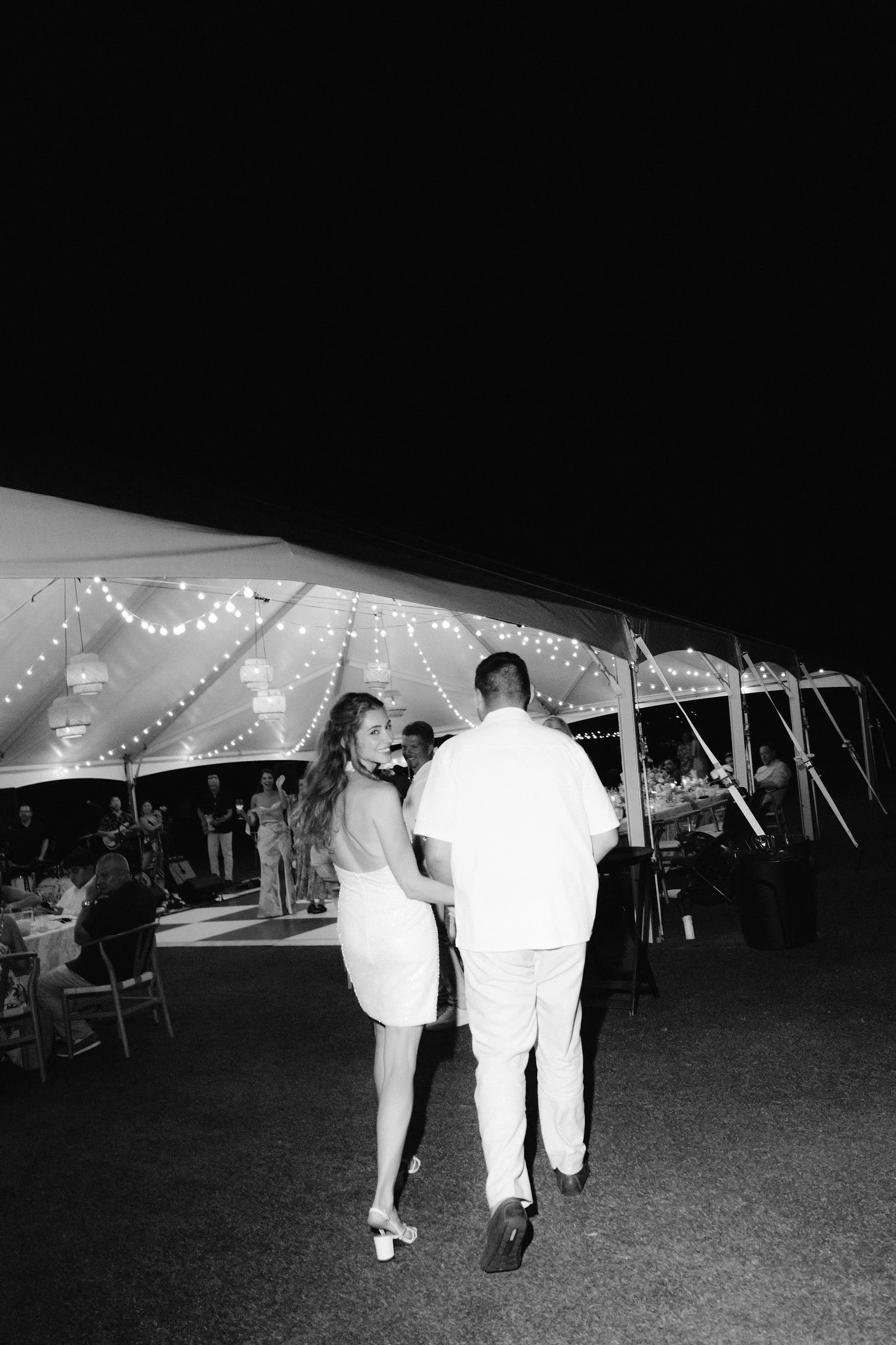 bride in her reception outfit looks back over her shoulder as her and groom walk into tent
