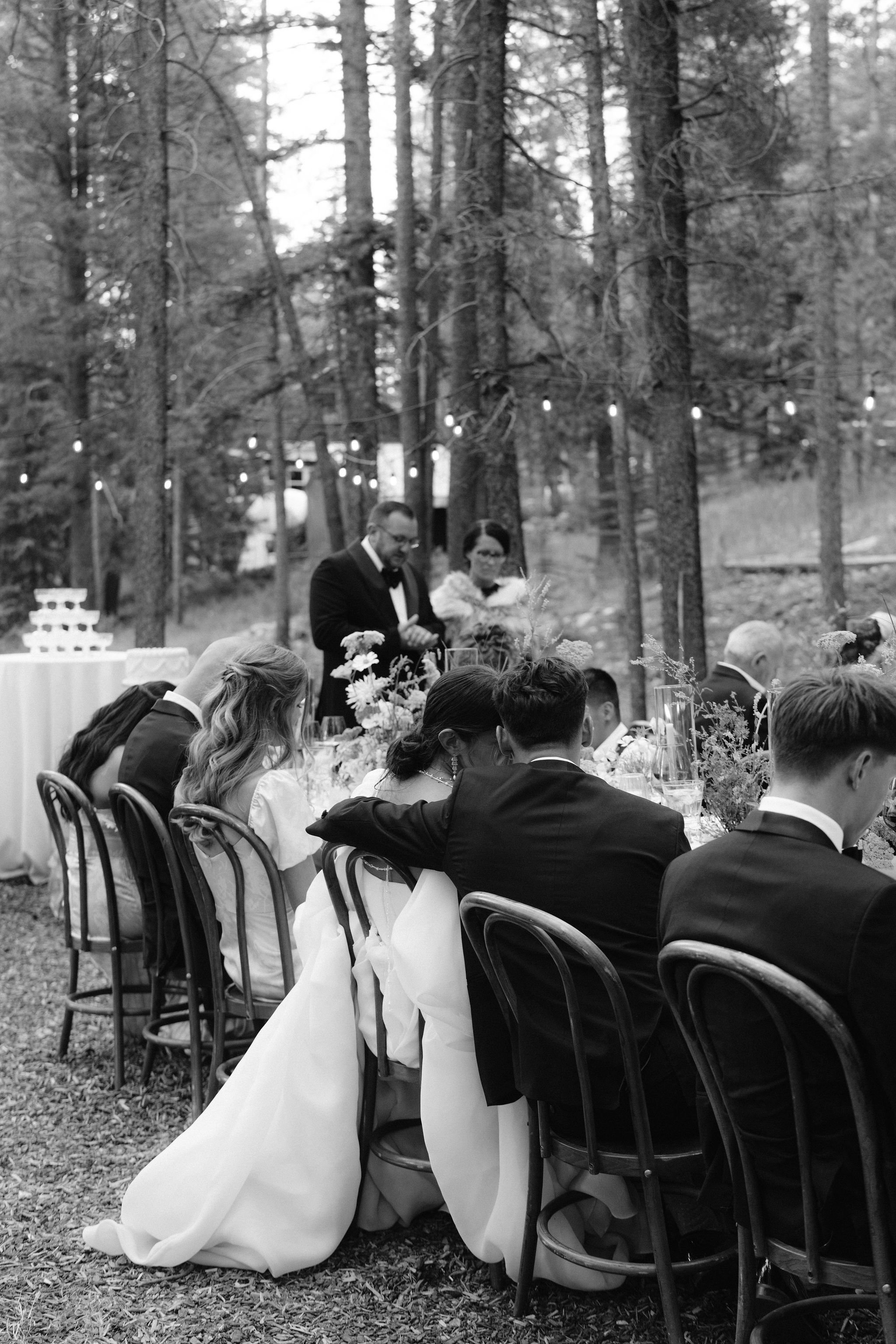 bride and groom seated for dinner and cuddle together as the parents of the bride pray over the wedding reception meal