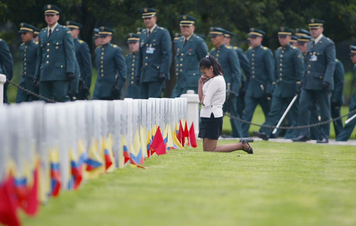 A woman cries as she kneels among the tombstones while army officers march at a military cemetery in Bogota, Colombia, 2003. Friends and relatives gathered at the military cemetery to honor soldiers who have been killed in fighting against rebels during the country's civil war.
