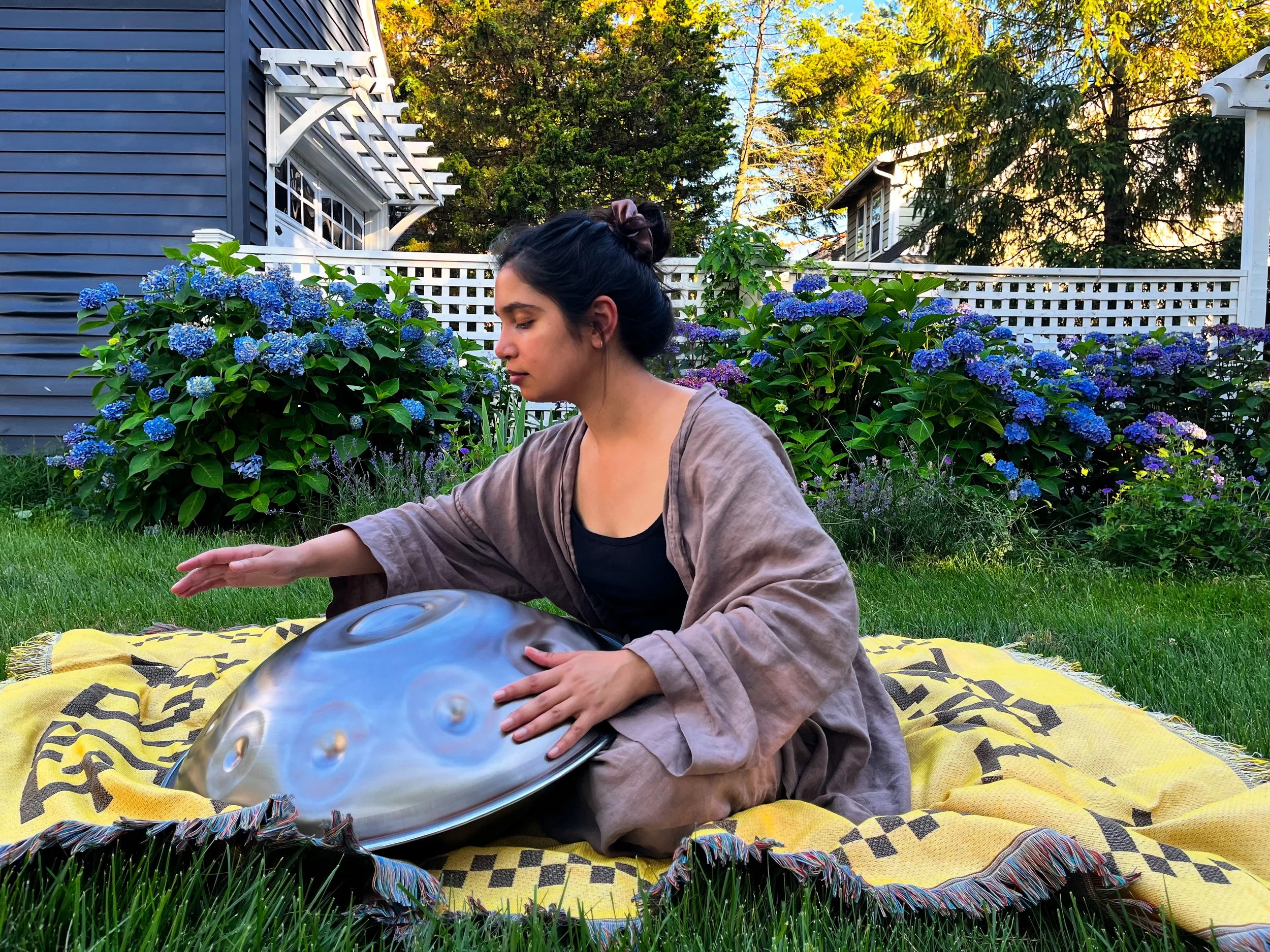 A woman with dark hair tied back, sitting outdoors on a yellow patterned blanket in front of green hydrangea bushes, with a blue house and trees in the background, playing a steel pan drum.