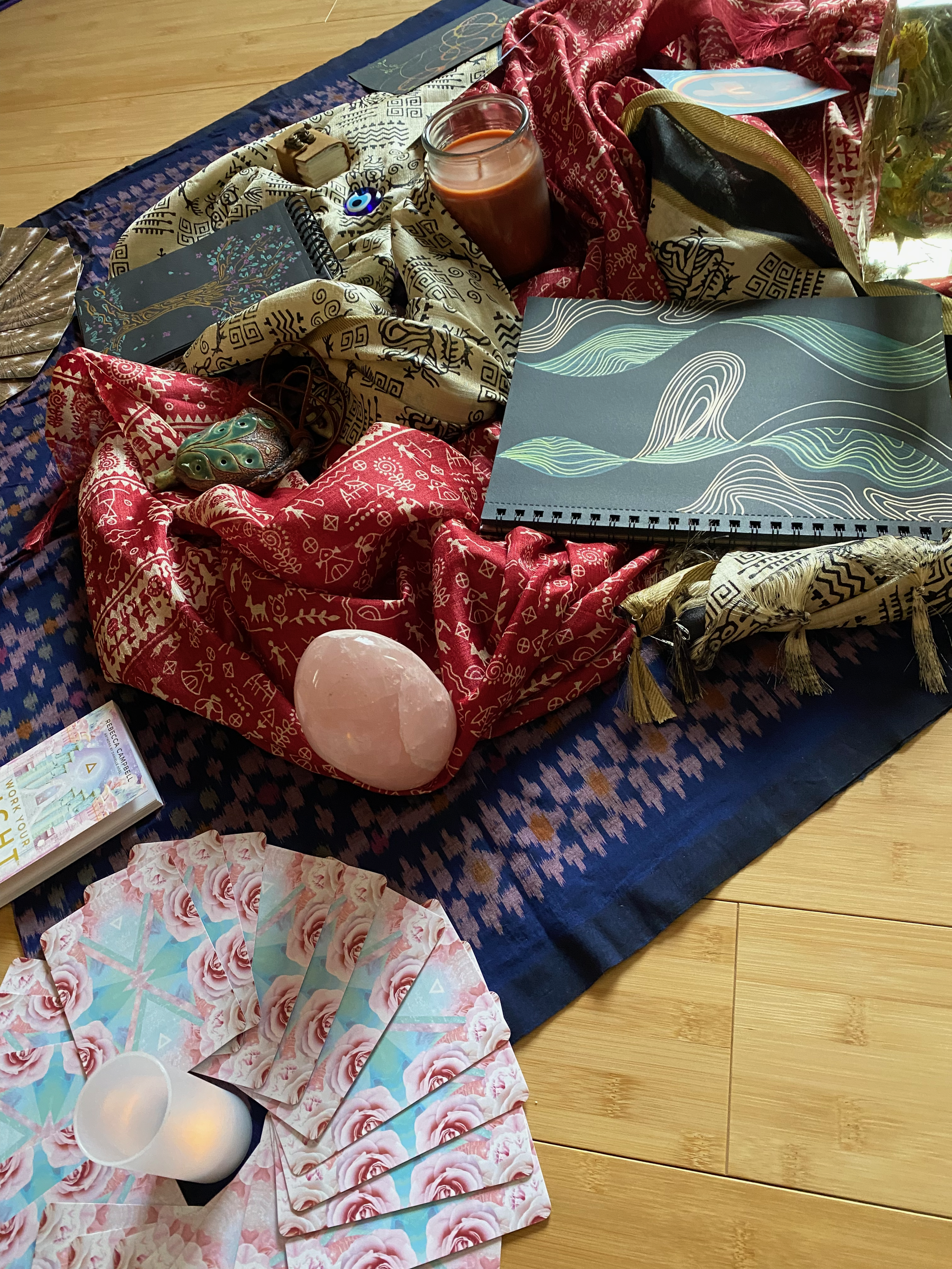 The image shows an arrangement of spiritual and artistic items on a wooden floor, including oracle cards, colorful fabrics, and various illustrations.