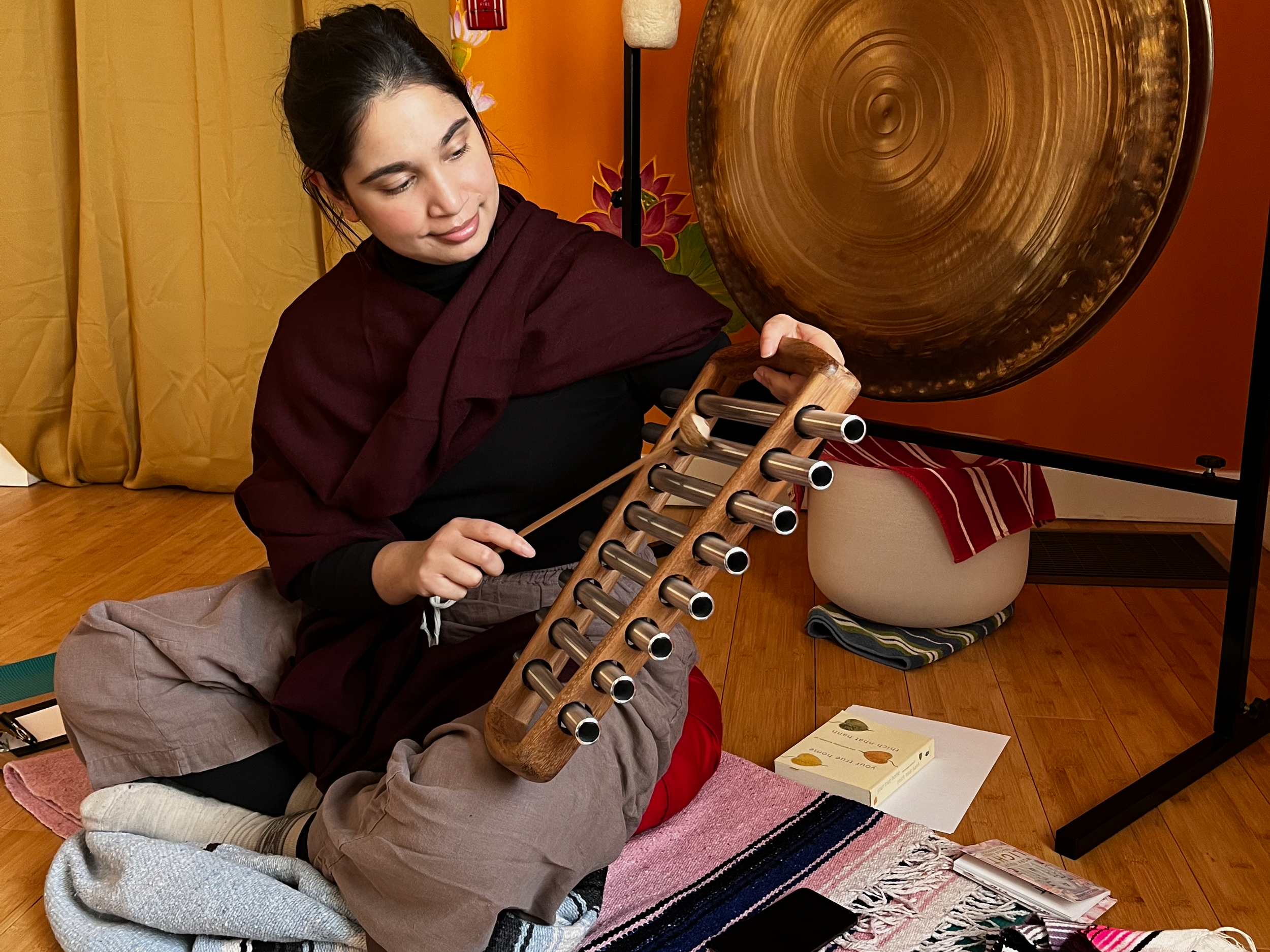 A woman in black clothing, with dark hair, sits cross-legged on a pink and white blanket on a wooden floor, playing a large gong with a mallet. To the right is a small table with a book and some magazines, and behind her is a yellow curtain and drying flower decoration on a stage.