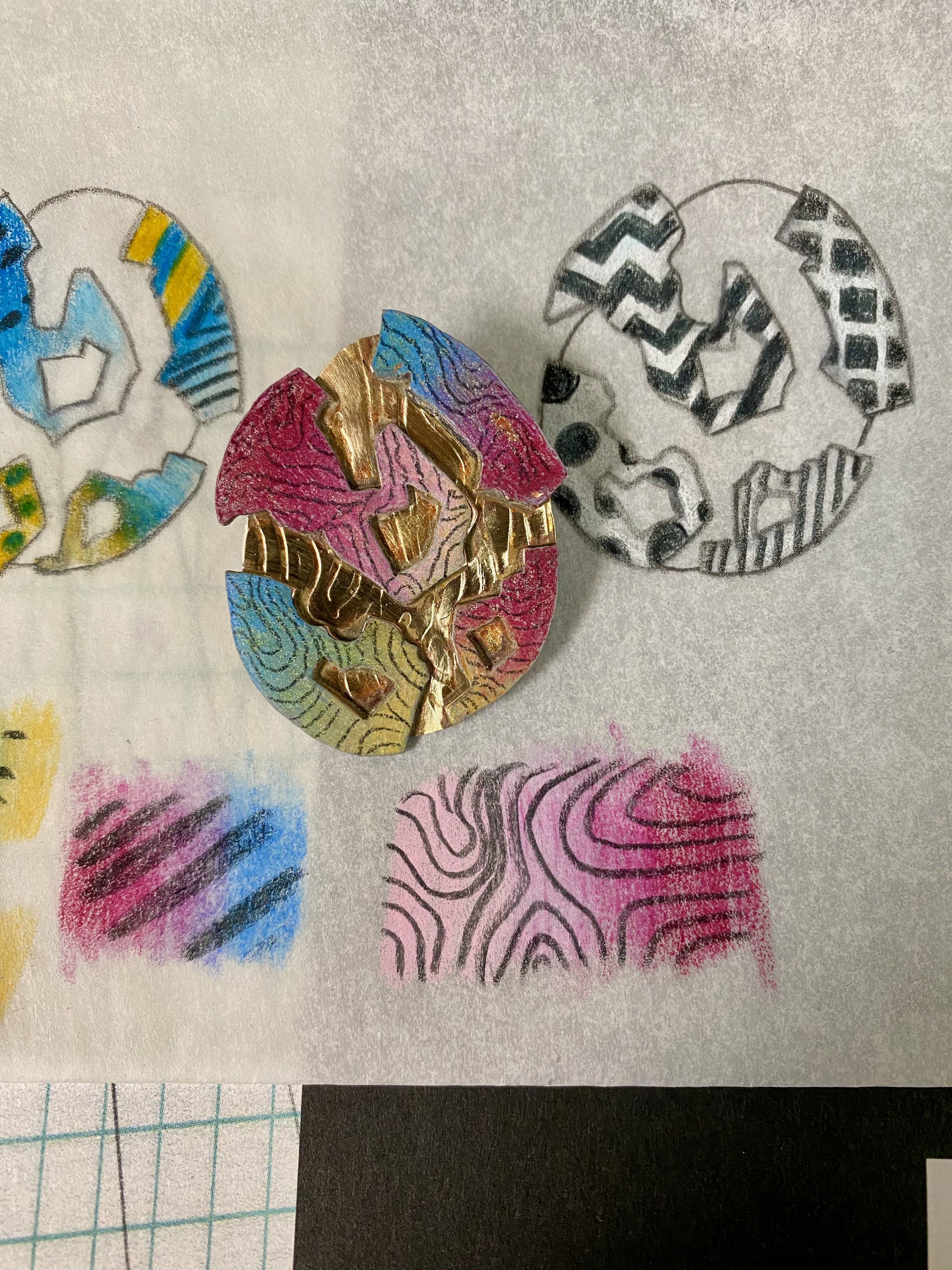 Colorful brass and copper pendant shaped like a cracked eggshell on a white background, surrounded by some colored pencil design samples featuring intricate black and white designs, and one with a rainbow pattern.