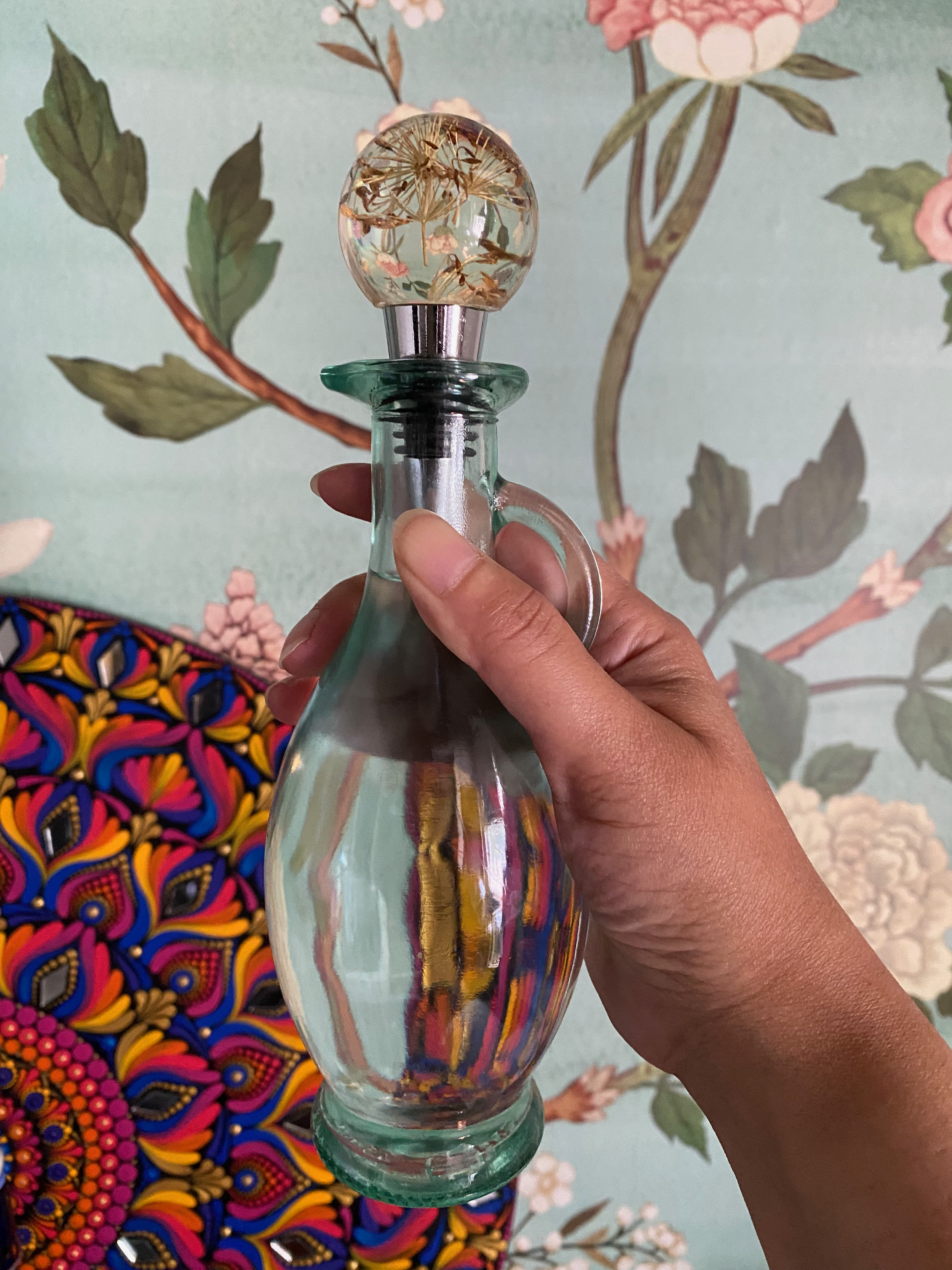 A hand holding a glass bottle of moon water with a spherical cap, against a background of colorful floral wallpaper and mandala.