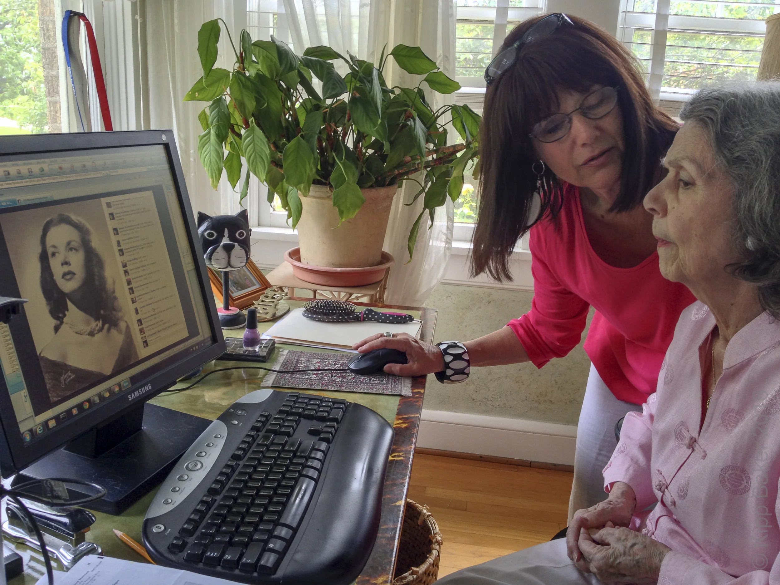 Author Bonnie Baker explains a Mother's Day Facebook post to her mother, artist, Olive Pemberton, shown in the image on the monitor at age 22. Copyright © Kipp Baker, All rights reserved.