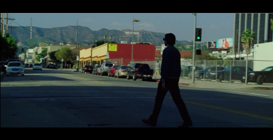 Screenshot from Nightcrawler of Lou Bloom crossing the street after he leaves the police station - the Hollywood sign in the far background quietly reminds us of Los Angeles as the other main character in the film.