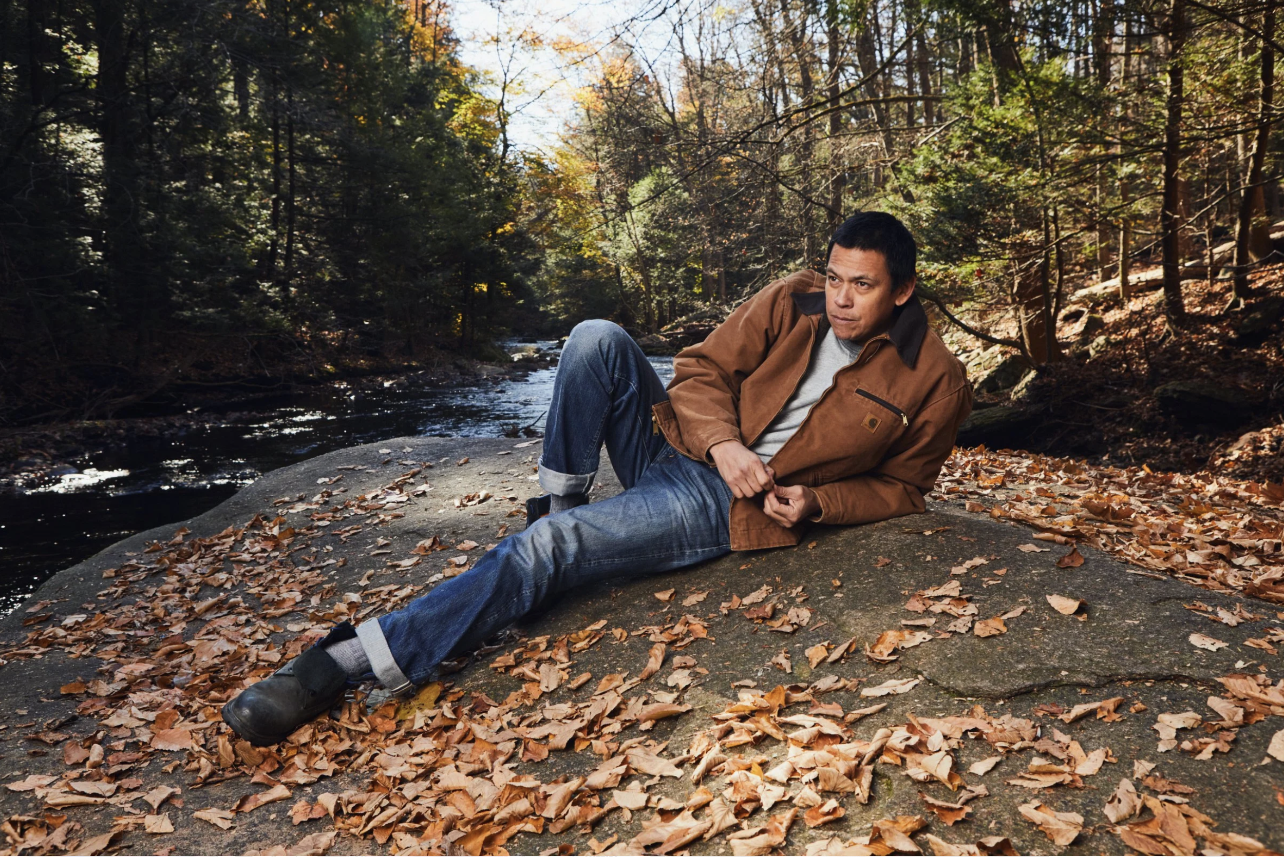 A man sitting on a large rock by a creek in a forest with autumn leaves falling and trees with colorful foliage.