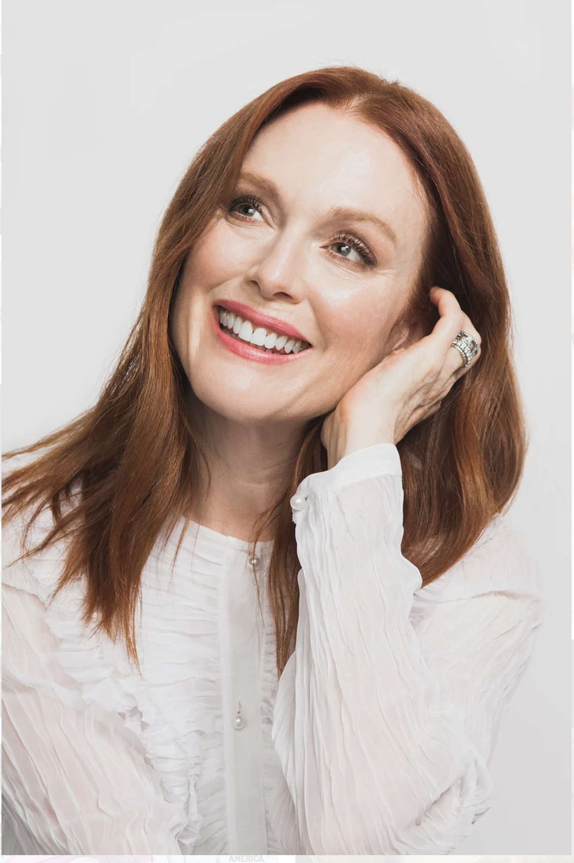 A woman with red hair smiling, wearing a white blouse, resting her head on her hand, against a plain light background.