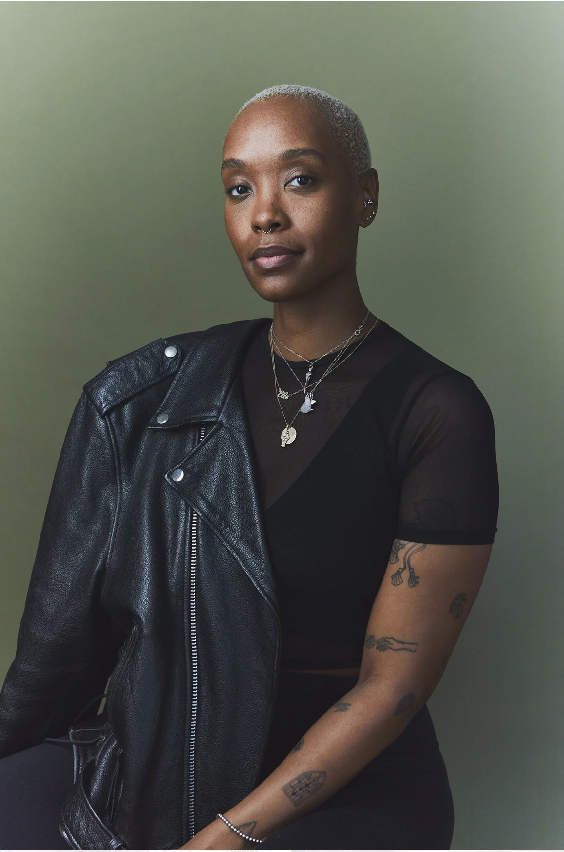 Portrait of a young woman with short blonde hair, wearing a black t-shirt with sheer sleeves, layered necklaces, and a black leather jacket over her shoulder, standing against a green background.
