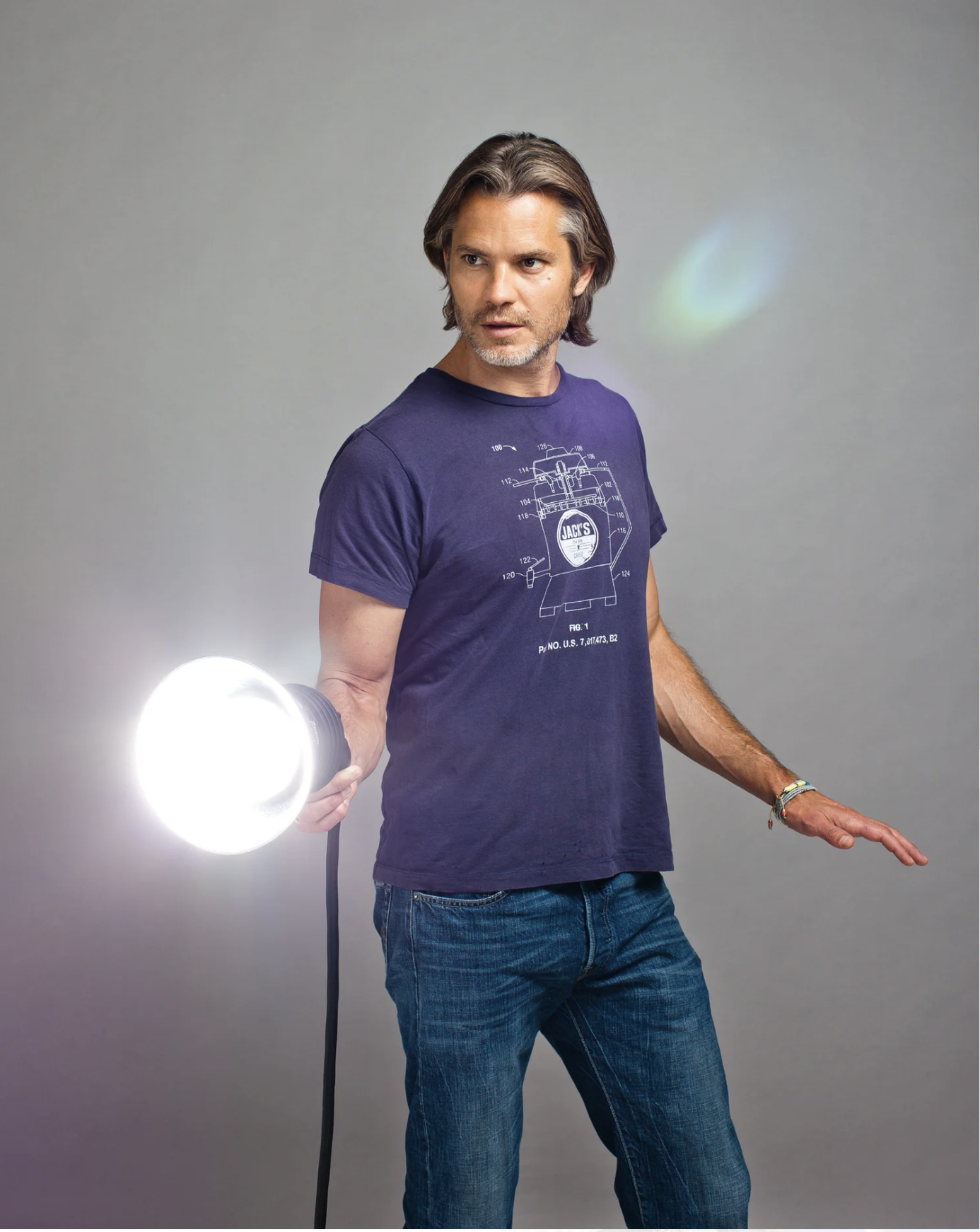 A middle-aged man with shoulder-length brown hair, a beard, wearing a navy t-shirt with technical drawings and text, and blue jeans, holding a bright light in his right hand, standing against a plain gray background.