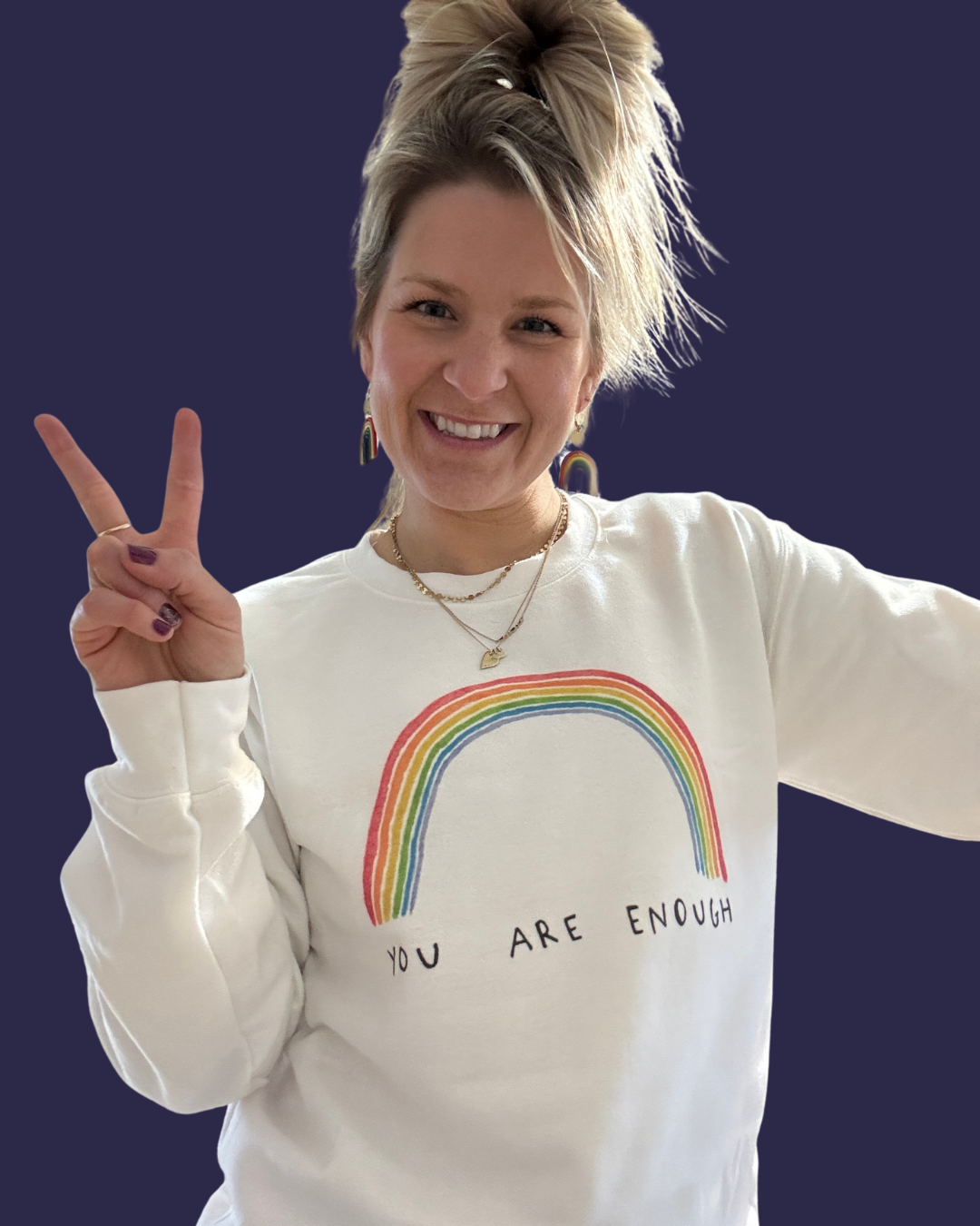 A smiling woman with blonde hair styled in a messy updo, wearing rainbow earrings, layered necklaces, and a white sweatshirt with a rainbow graphic and the words 'YOU ARE ENOUGH.' She is holding up a peace sign with her right hand and against a dark background.