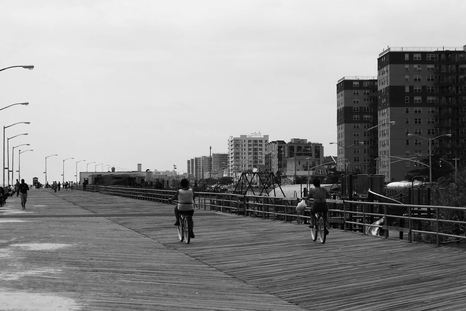 A black-and-white photo of a seaside boardwalk with people walking and cycling, tall buildings in the background, and street lamps lining the walkway.