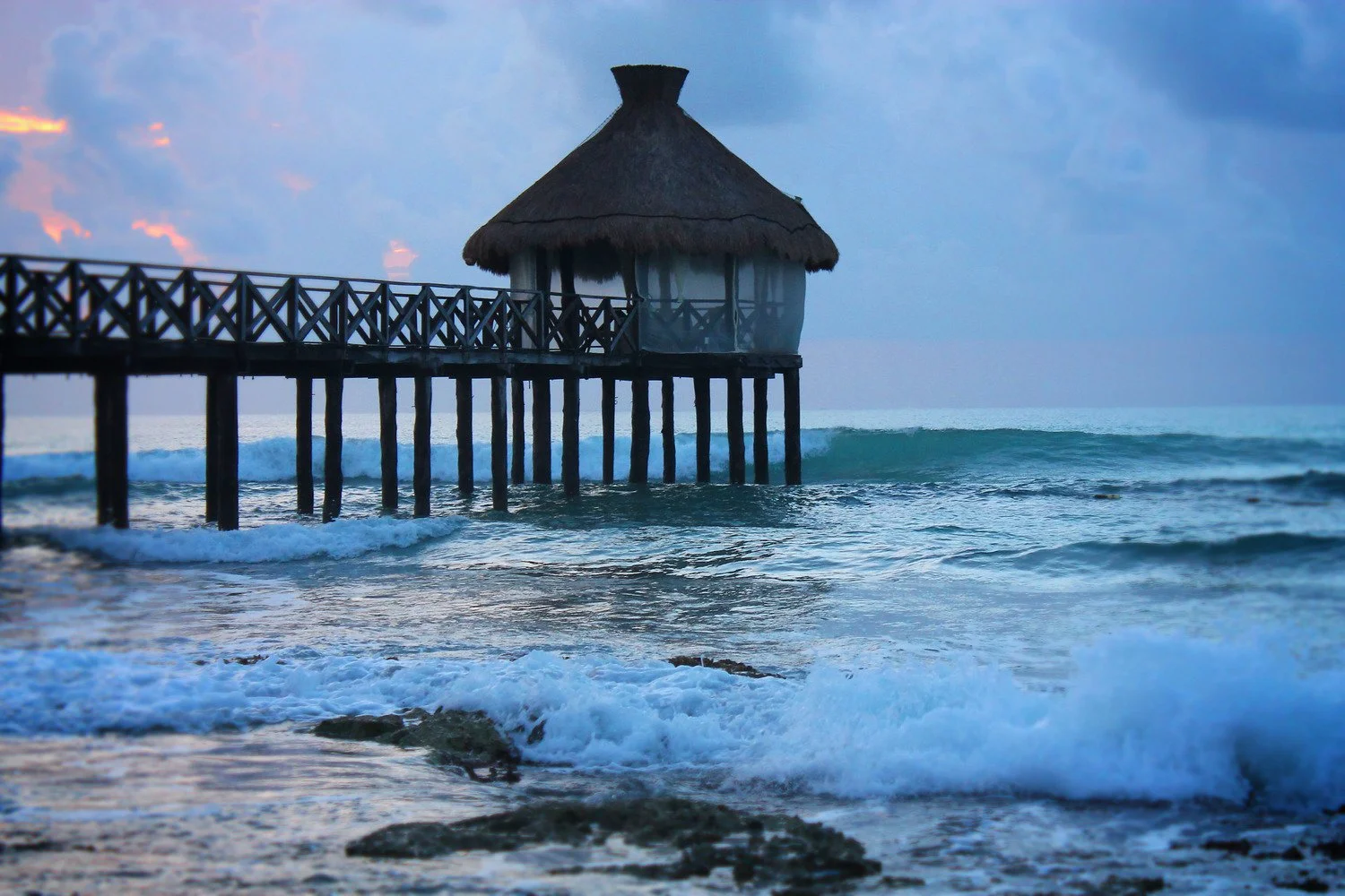 A wooden pier extending into the ocean with a thatched-roof hut at the end, waves crashing against the rocks, under a cloudy sky at dusk.