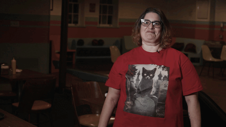 A woman with glasses and shoulder-length brown hair stands inside a dimly lit room, wearing a red t-shirt with a black cat on a gravestone printed on the front.