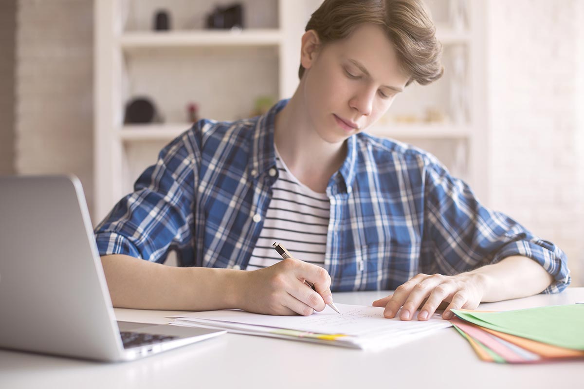 High school student drafts a Common App essay in a notebook while working at a laptop