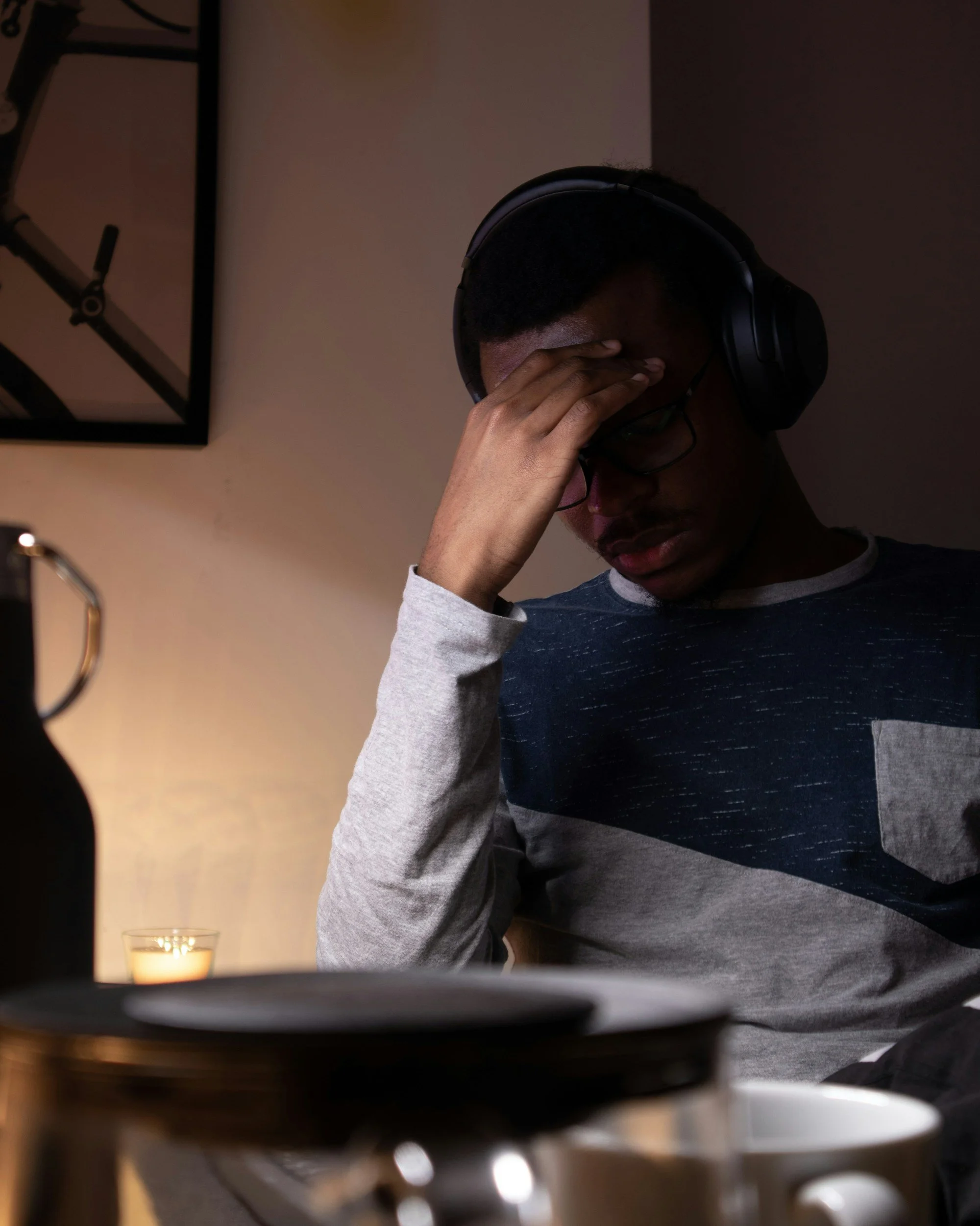 African Marian man with glasses and headphones with head in one hand in a room with soft lighting looking tired and overwhelmed