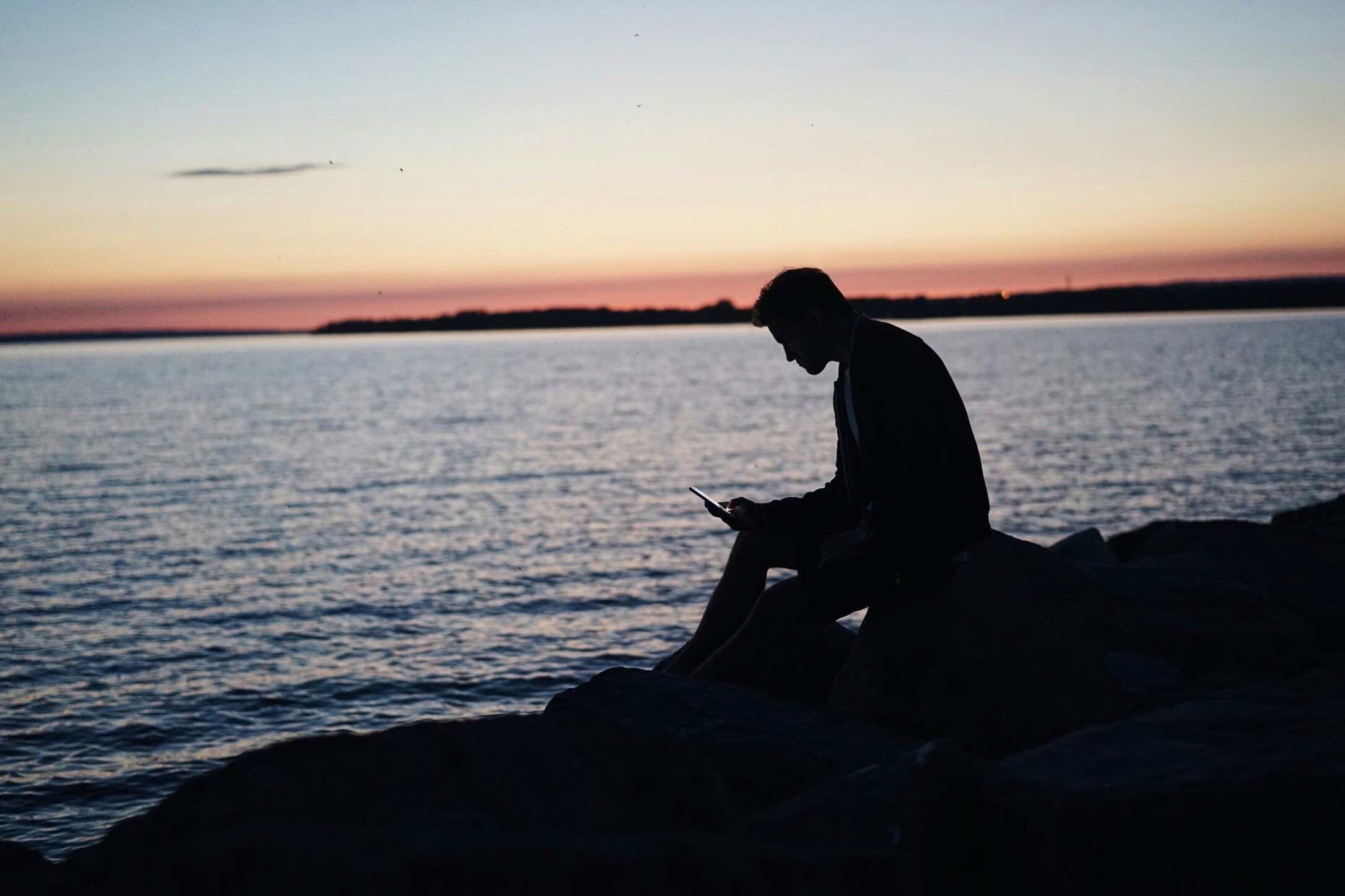 man sitting on a rocky  beach at sunset looking at his phone