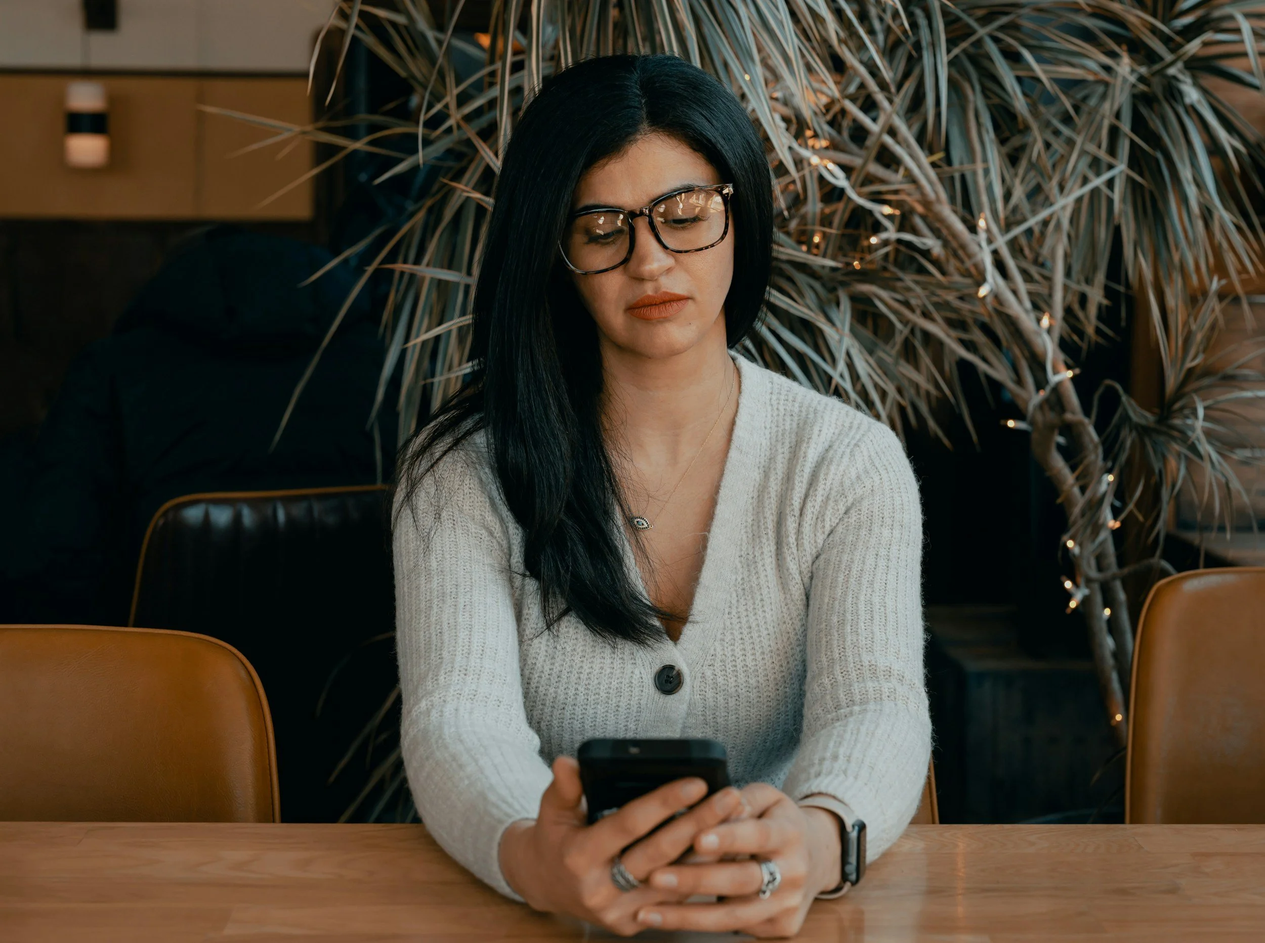 woman with dark hair and glasses sitting at a table looking at her phone
