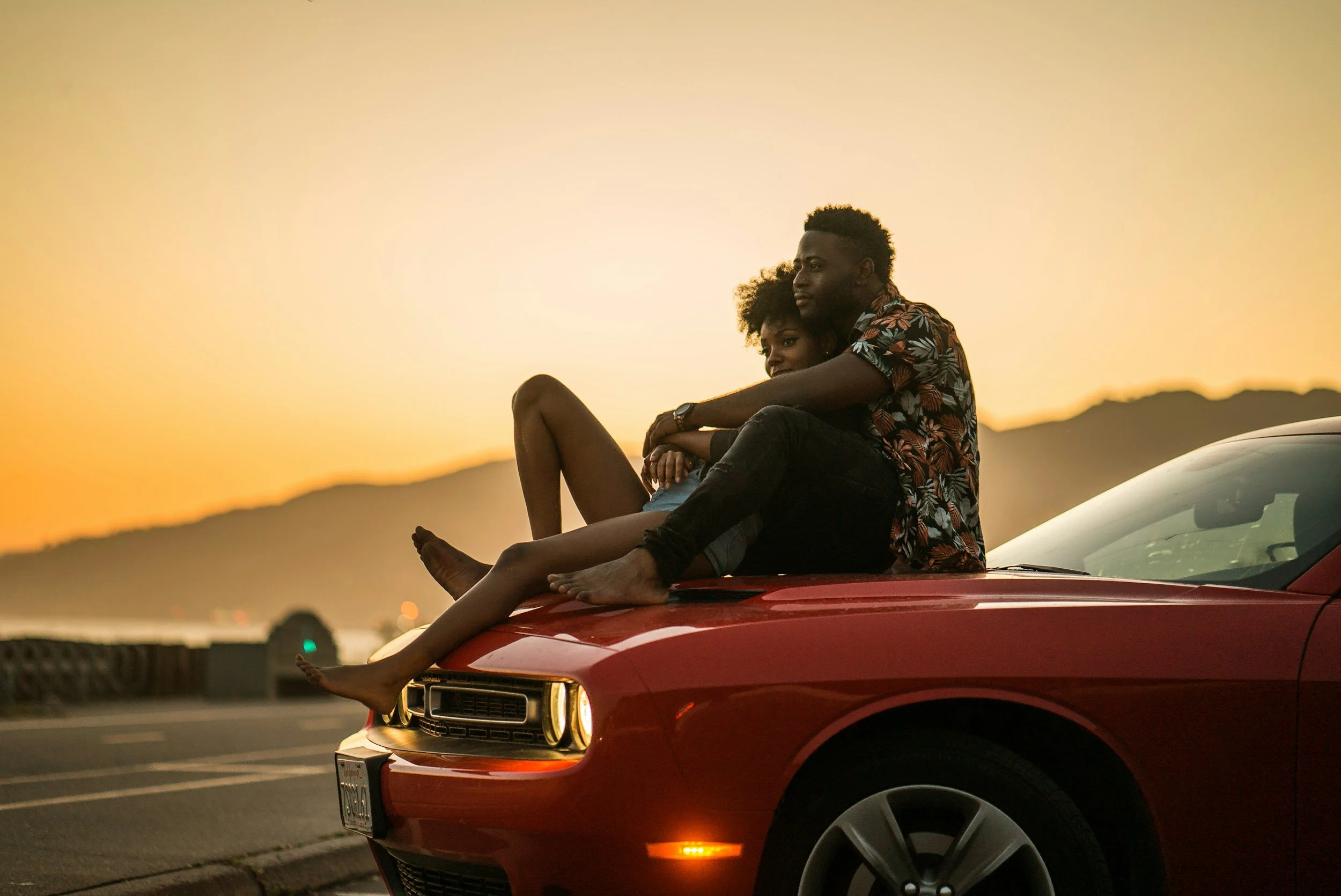 black couples sitting on the hood of a red car watching the sunset at the beach