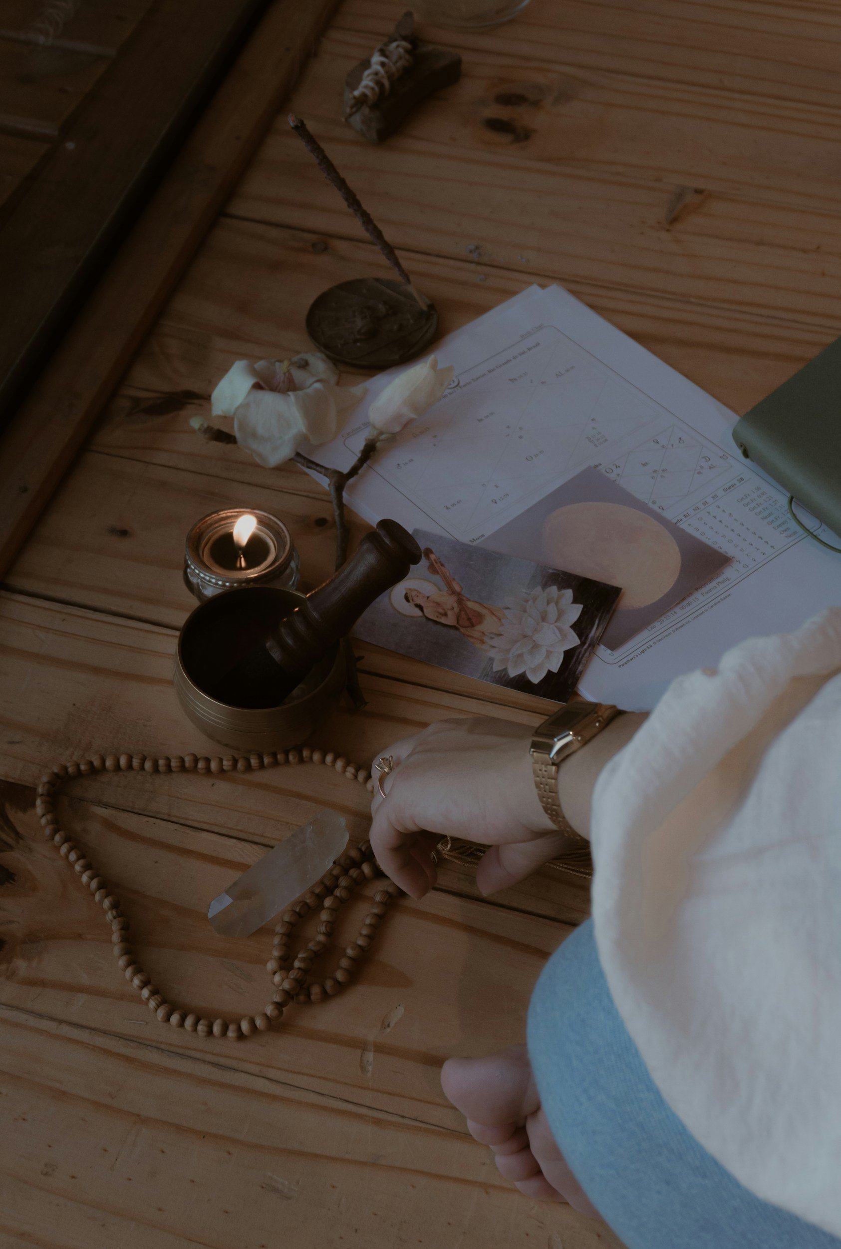 A person wearing a white long sleeve shirt and a silver watch is holding a clear quartz crystal in their hand. The surface in front of them has various objects including a lit candle, a string of wooden prayer beads, a framed picture with a lotus flower and moon, some papers with handwritten text, a small object with a branch and rocks on top, and a menu or booklet. The scene is on a wooden surface with a wood-paneled wall background.