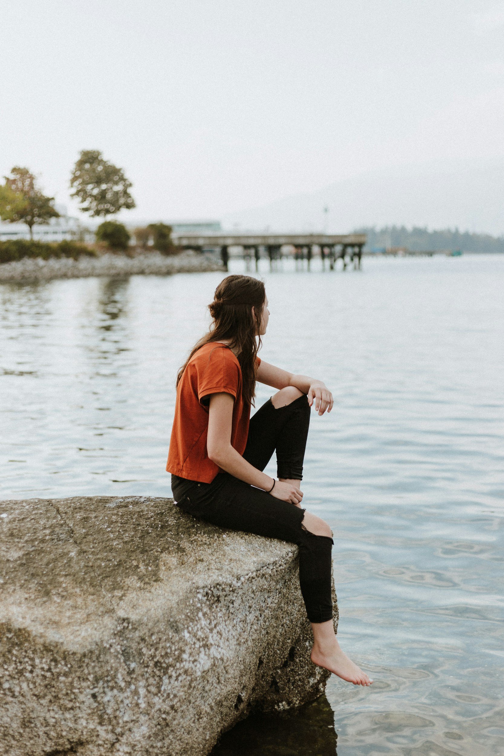 woman with dark hair, orange t-shirt and black skinny jeans with the knees torn sitting on the edge of a rock with her feet dangling over water looking to her left