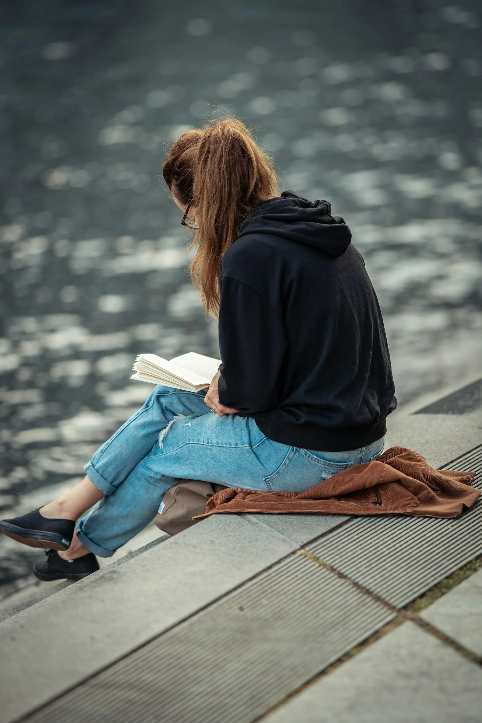 woman in a black sweatshirt and jeans reading a book on cement steps overlooking water