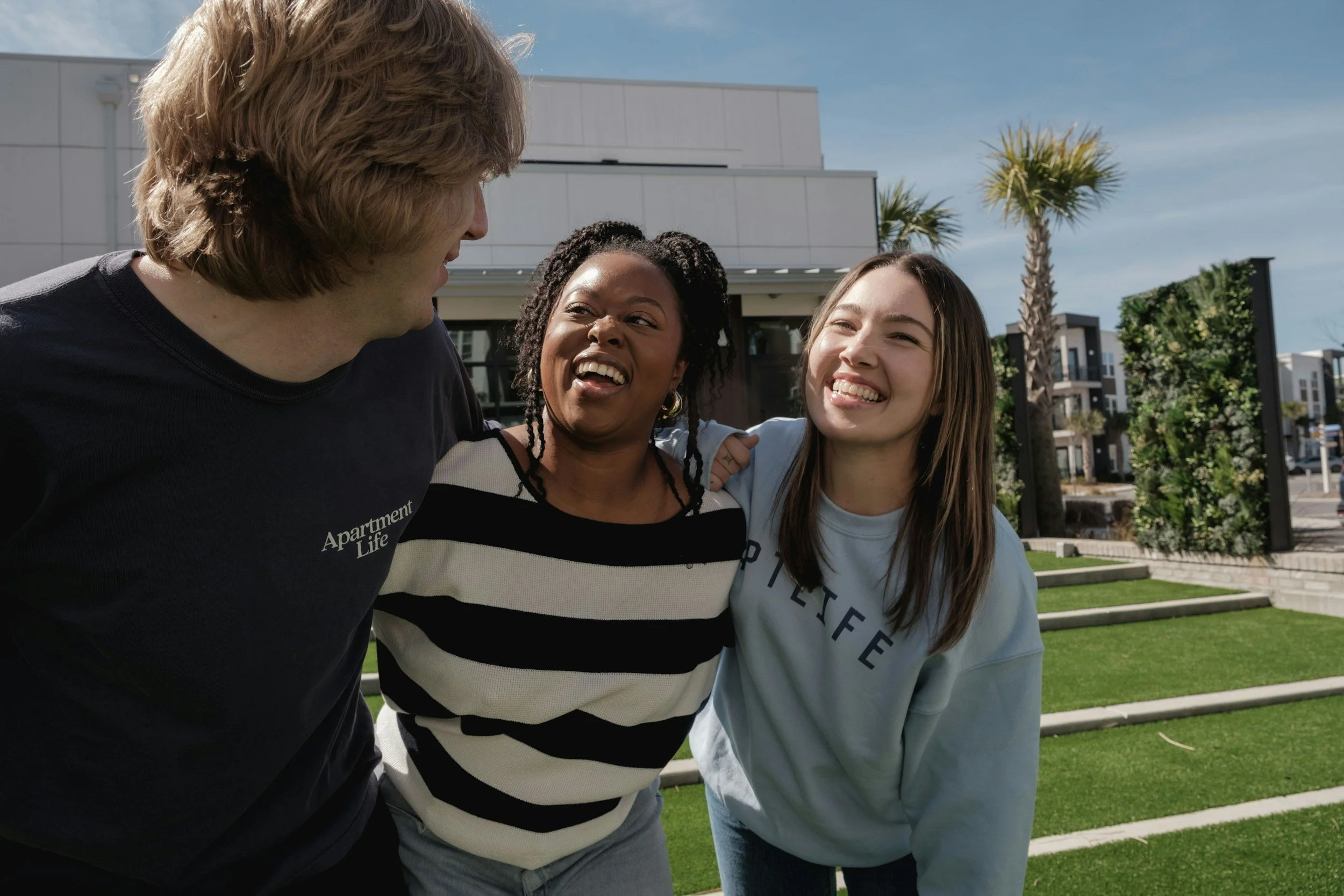 a group of three people standing shoulder to shoulder all smiling and lookout at one another outside infant of a white building with grass signifying community connections