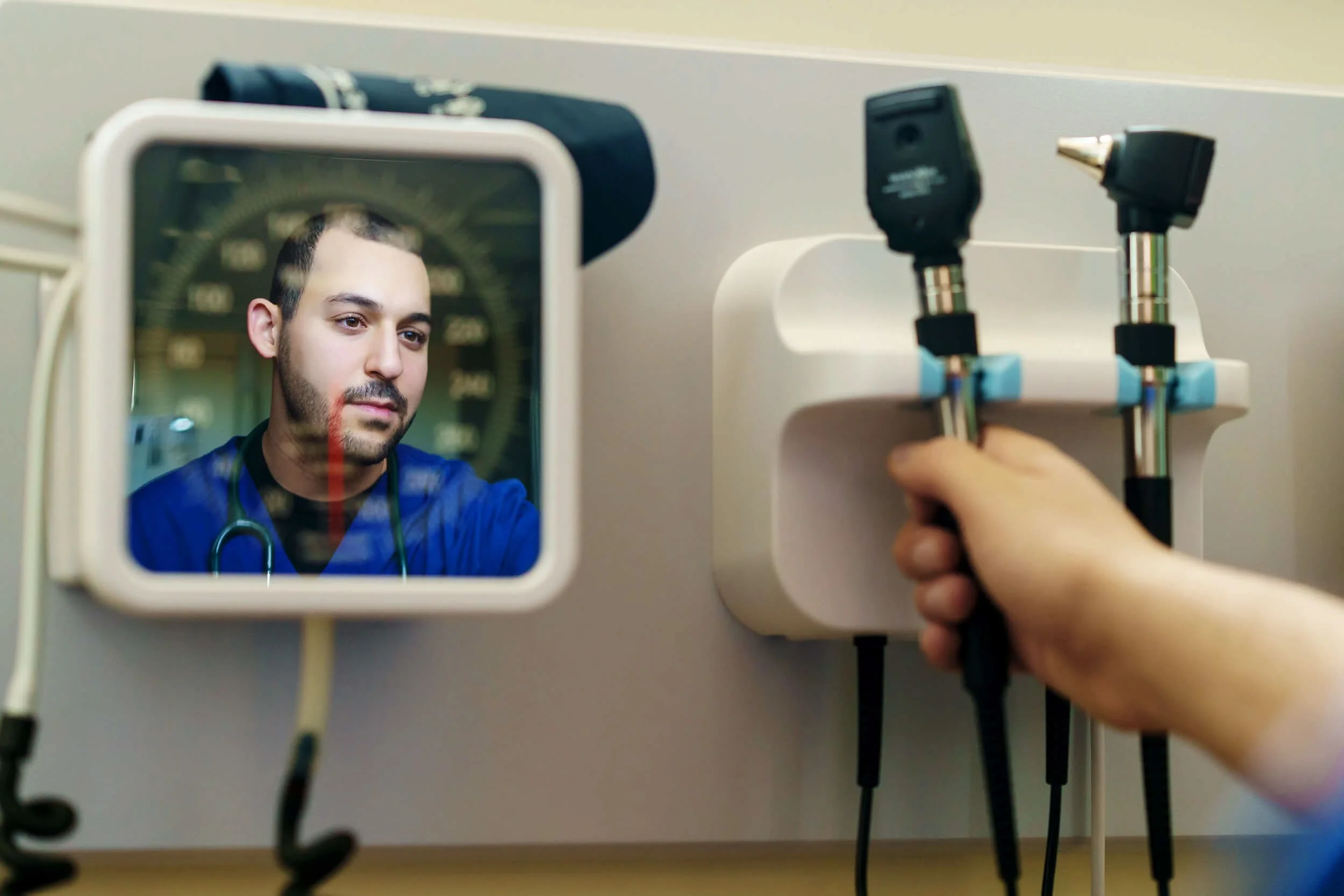 A healthcare worker's reflection seen in a mirror with a stethoscope around his neck, as a hand plugs in medical equipment into a wall socket.