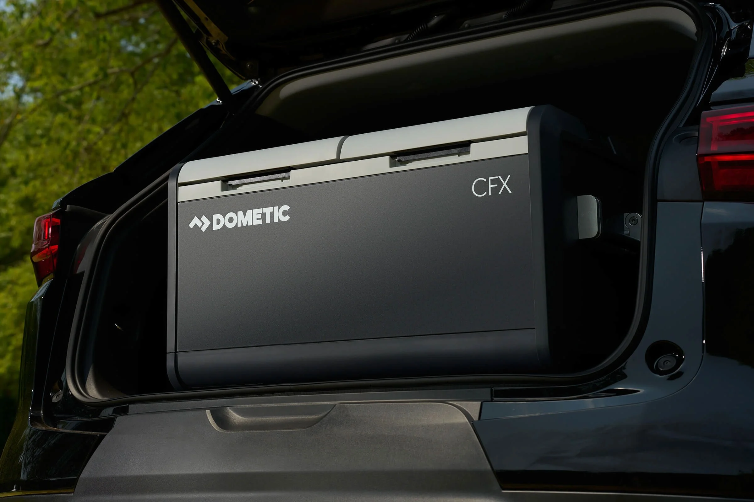Electric vehicle trunk open revealing a Dometic CFX portable refrigerator in black, with a logo and model name, in a vehicle outdoors with green trees in the background.