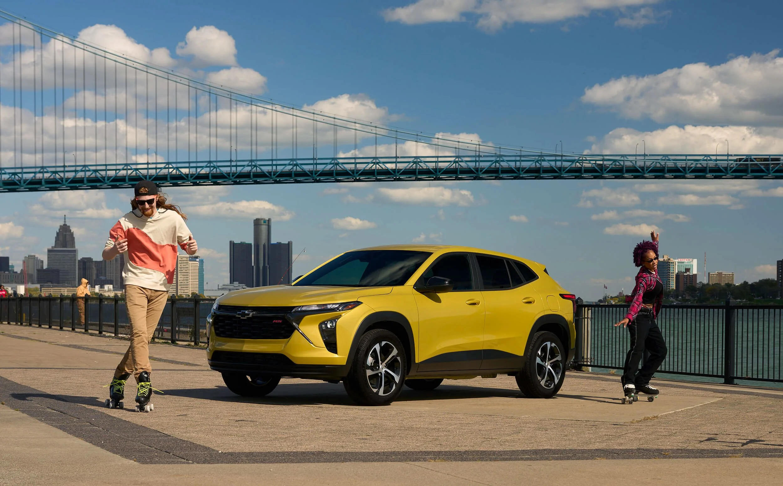 Two people roller skating next to a yellow SUV on a waterfront promenade with city skyline and a bridge in the background on a sunny day.