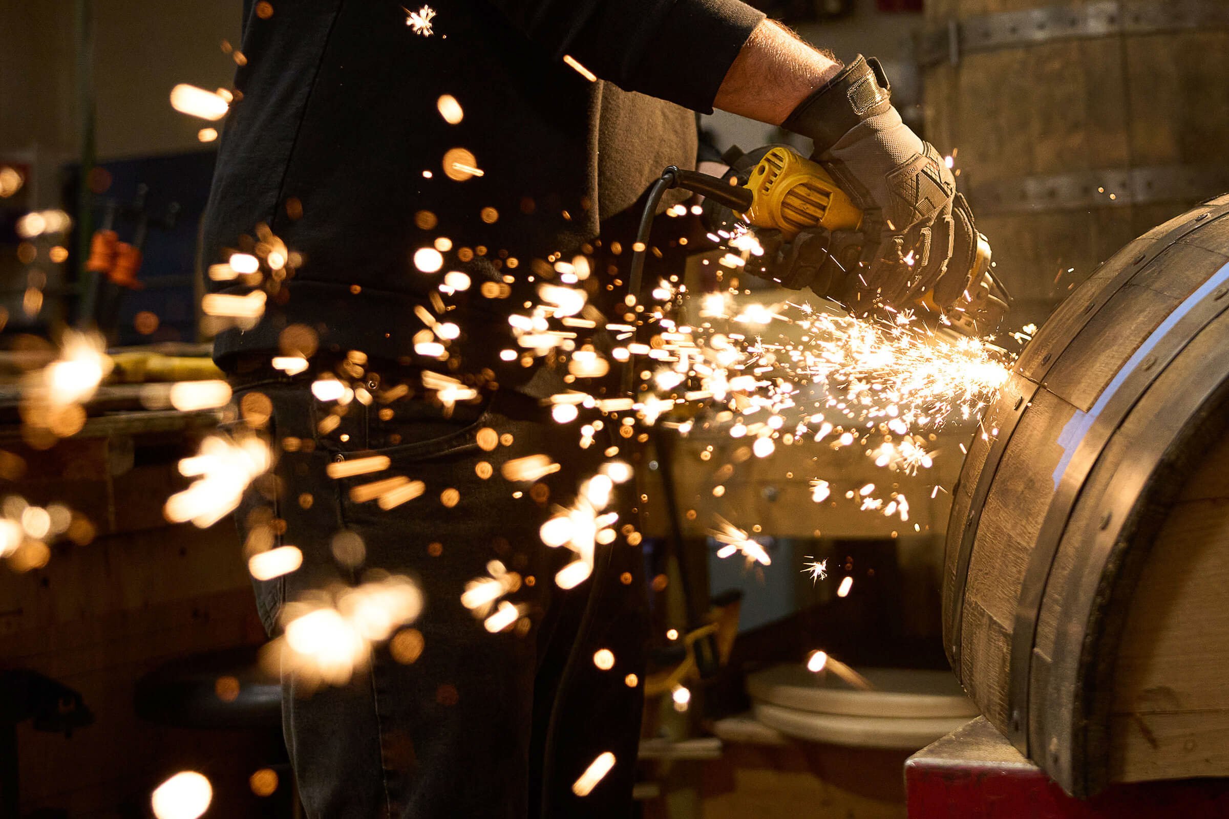 A worker wearing gloves and using a power tool to cut or grind a large wooden barrel, with sparks flying.