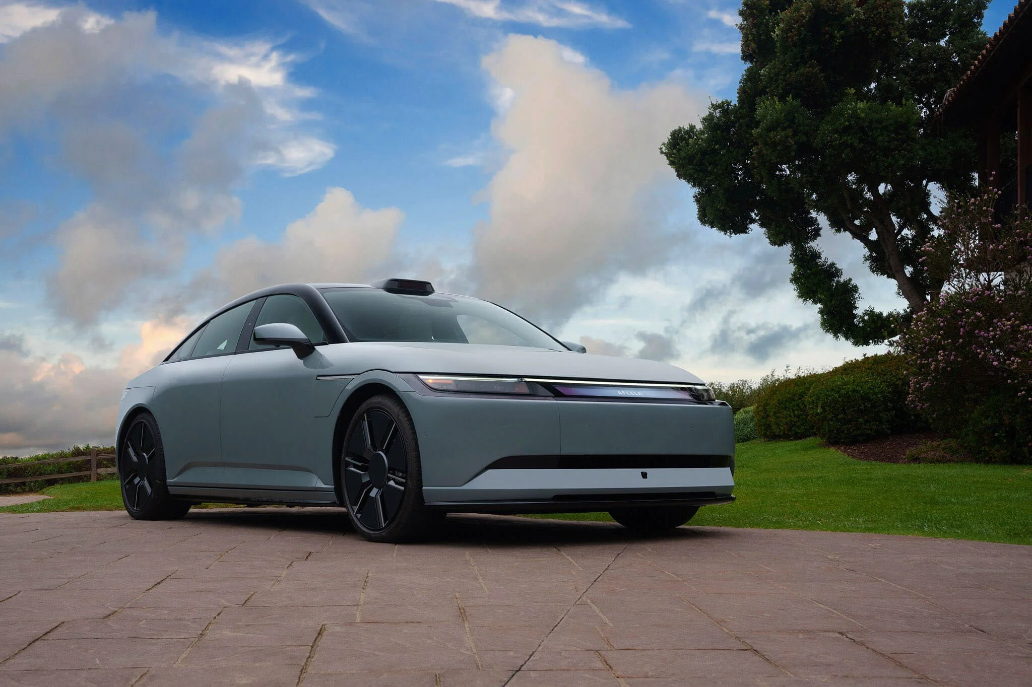 A sleek, modern silver electric vehicle parked on a paved driveway with grass and bushes on the side. The sky above is partly cloudy with a mix of blue and white clouds.