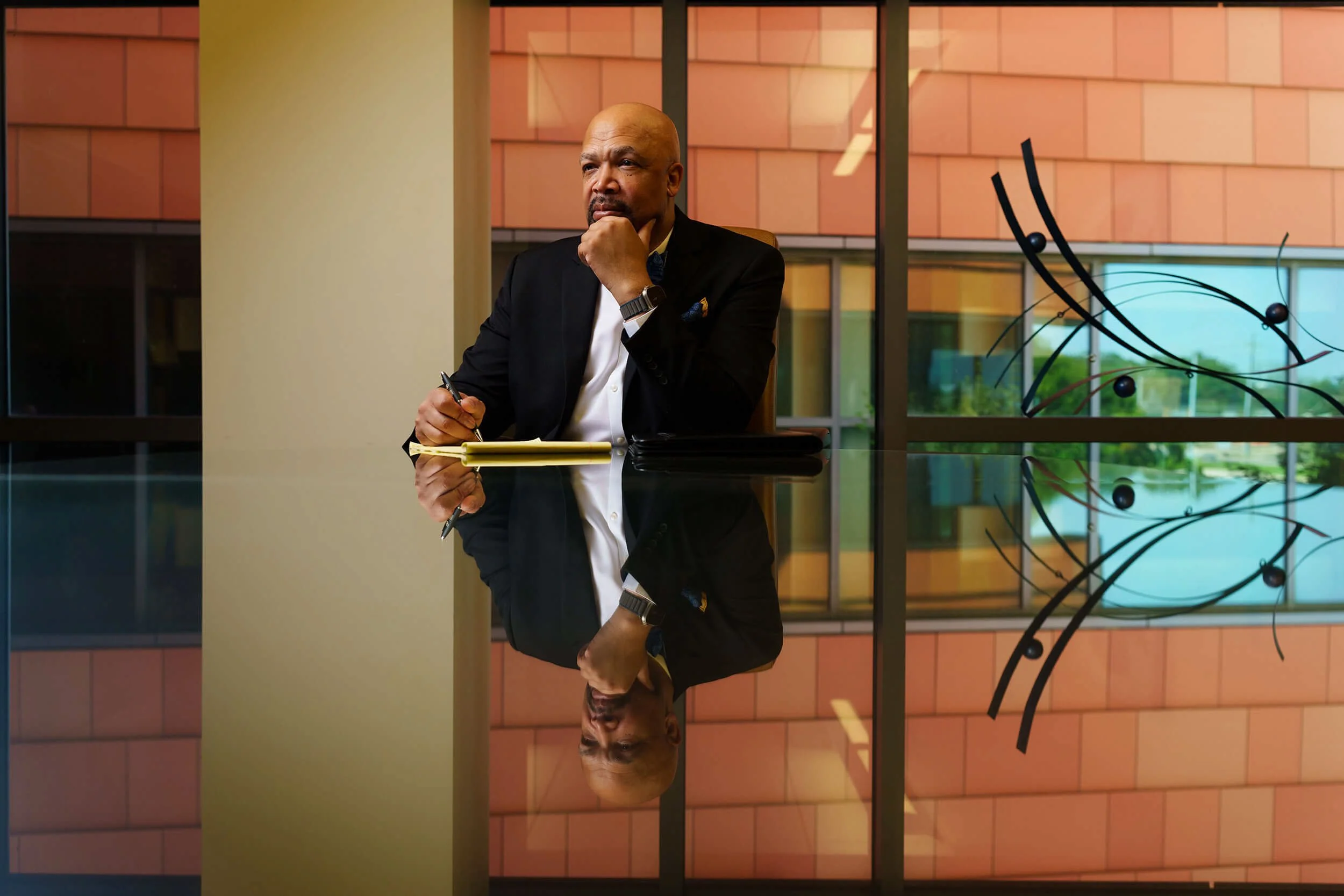 A man dressed in a black suit and white shirt sitting at a conference table, holding a pen and looking thoughtfully, with a notebook open in front of him. The table has a highly reflective surface, creating a mirror image of the man and the backgroun