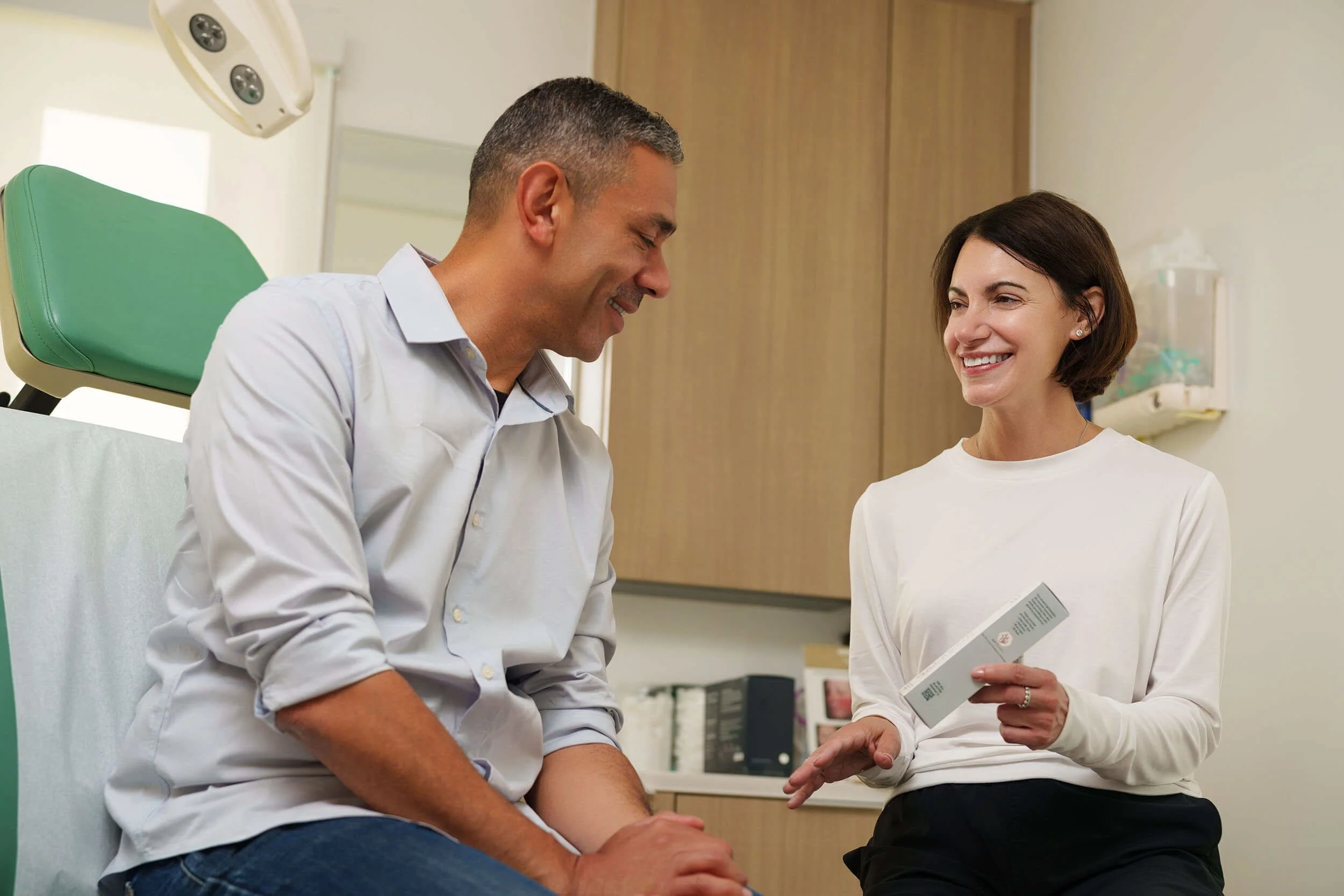 A woman in a white long sleeve shirt sitting in a medical office, smiling and talking to a man in a gray shirt sitting on an exam table.