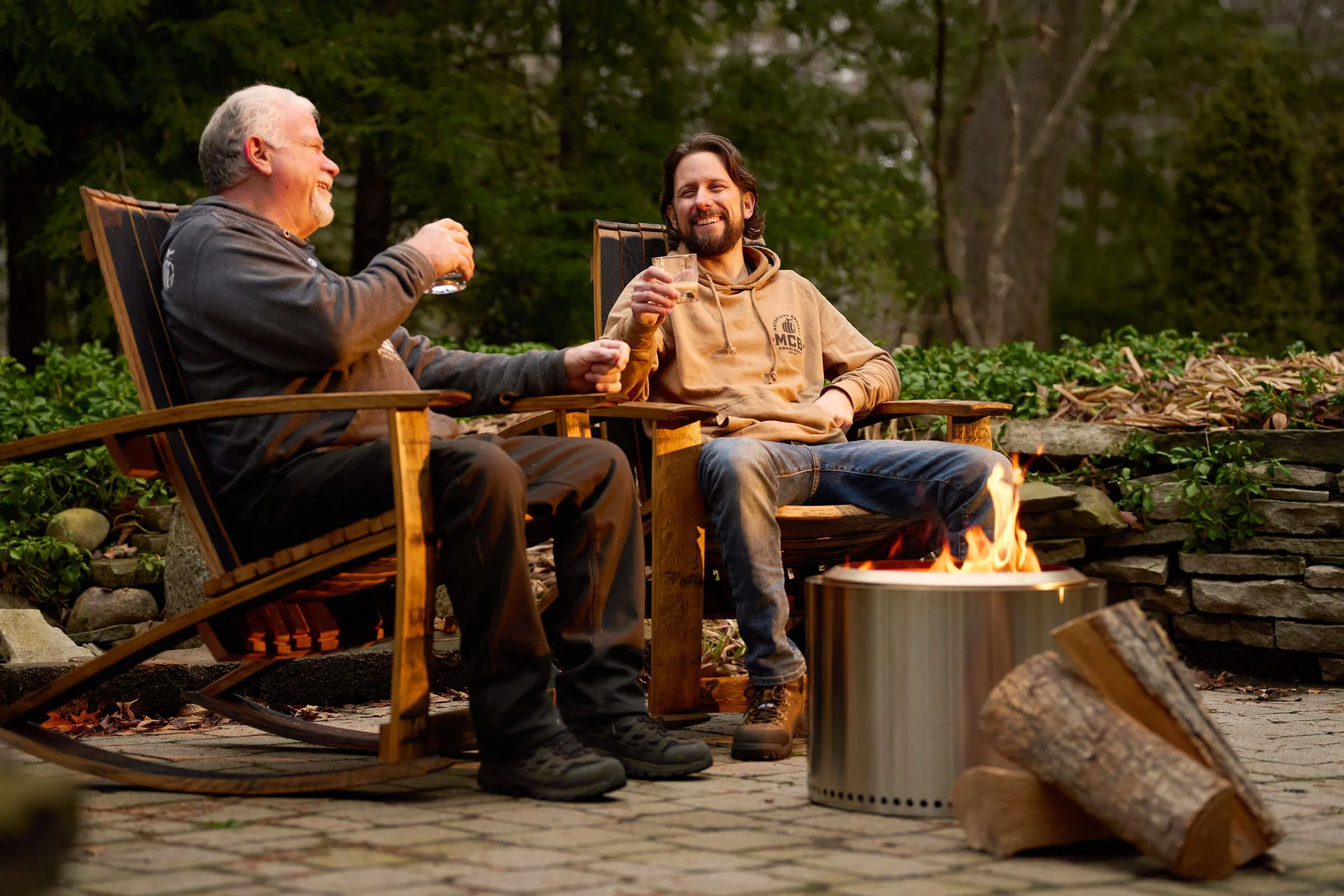 Two men sitting in wooden chairs outdoors near a fire pit, smiling and drinking beverages, surrounded by trees and greenery.