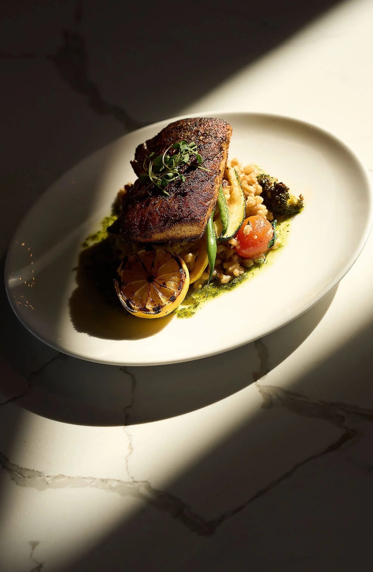 A plated gourmet dish featuring a seared piece of fish with herbs, served with grilled lemon, roasted vegetables, and rice, on a white oval plate with dramatic lighting.