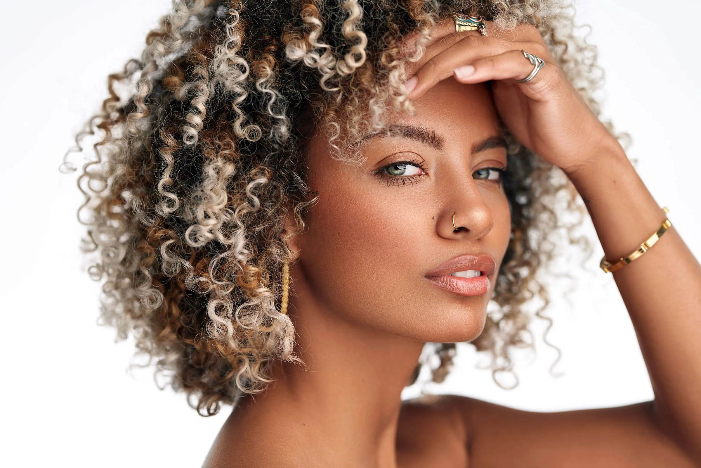 A woman with curly, blond and black hair, wearing gold earrings, a nose ring, and gold jewelry, posing with her hand on her forehead against a white background.
