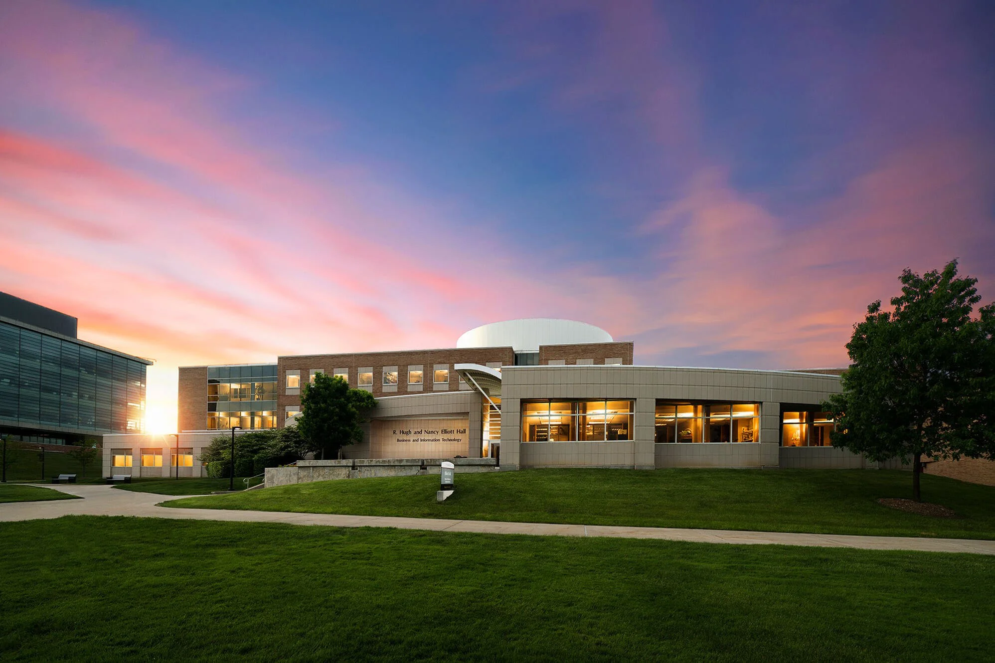 The R. Hugh and Nancy Elliott Hall building on a college campus at sunset, with a colorful sky and green lawn in the foreground.