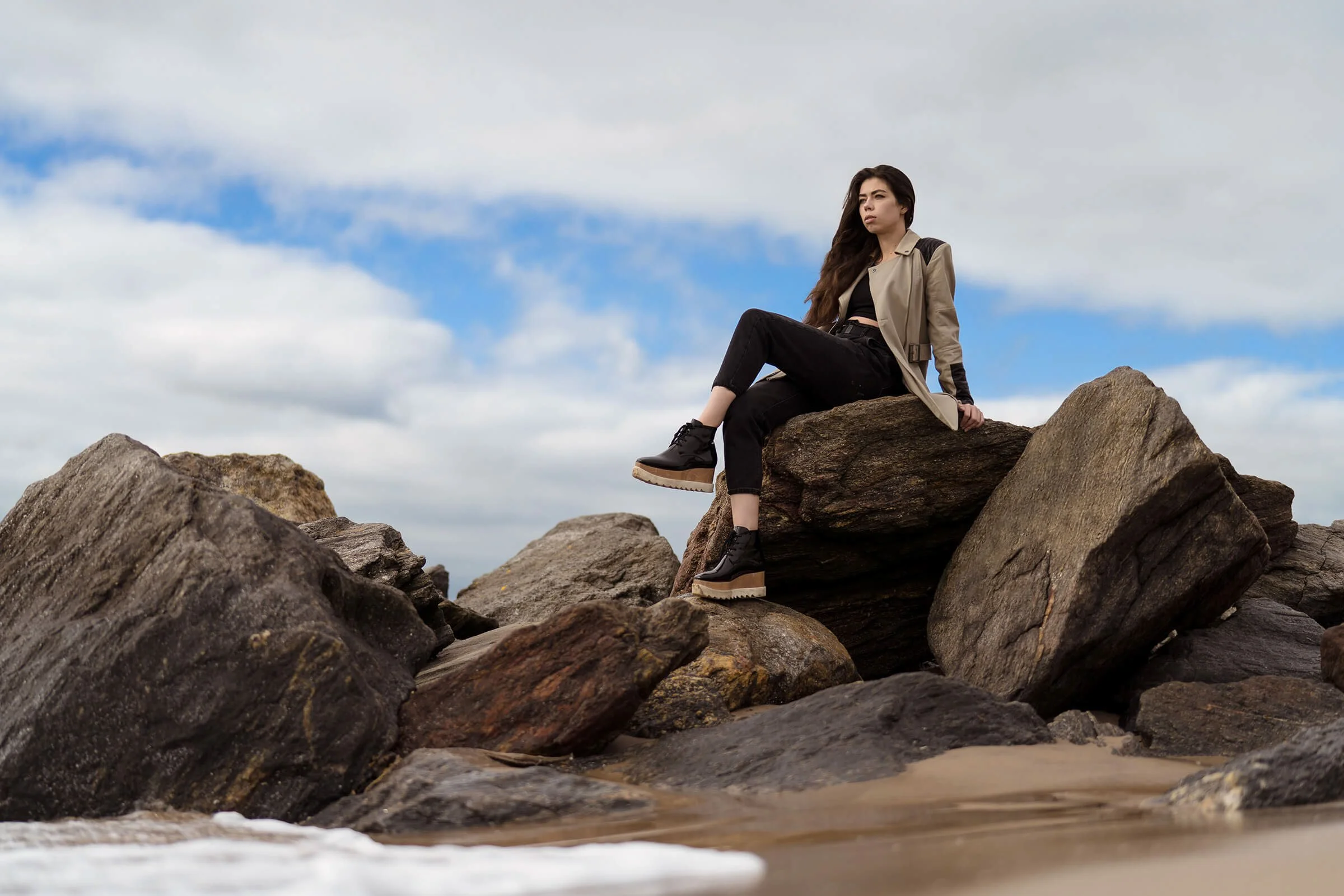A woman sitting on large rocks at the beach with a cloudy sky in the background.
