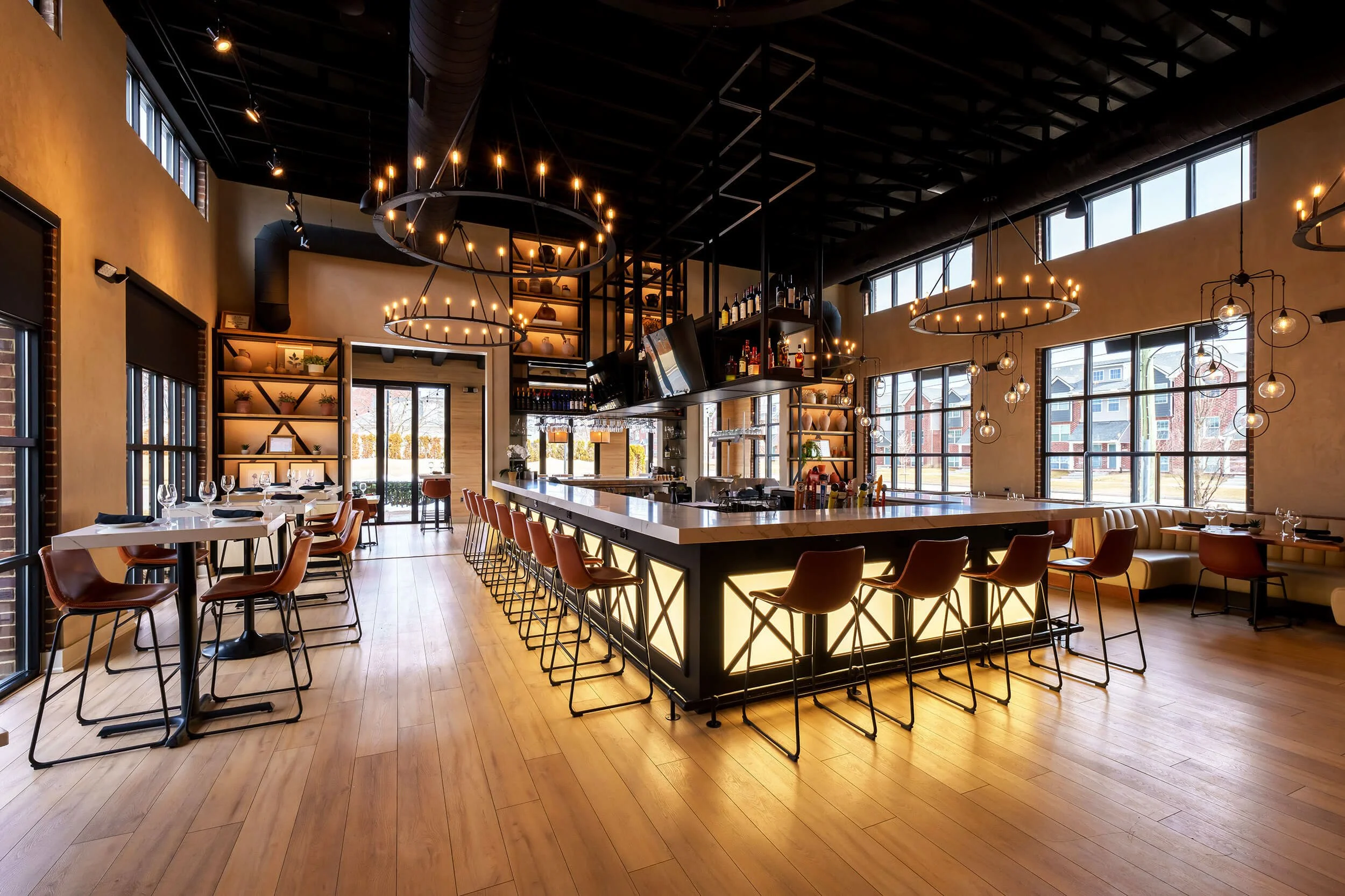 An upscale restaurant interior with a long illuminated marble bar, high-top counters, and seating. Large windows along the right wall let in natural light, with additional pendant and chandelier lighting fixtures hanging from the black-painted ceilin