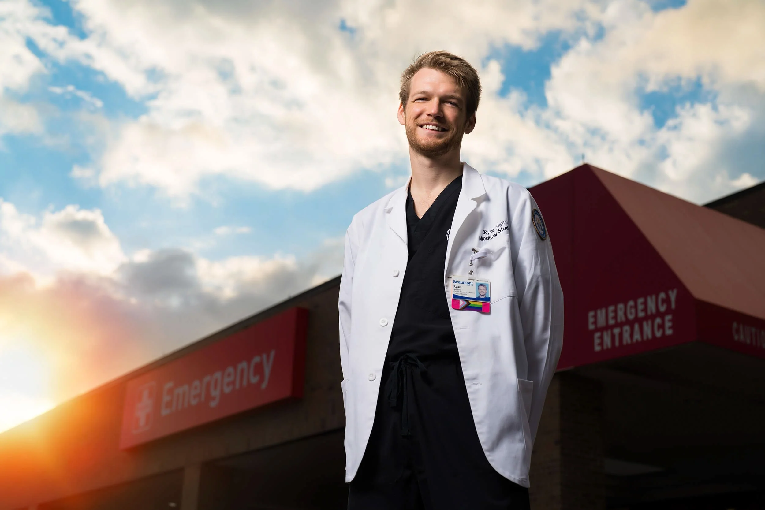 Smiling male doctor standing outside emergency room at sunset, wearing a white coat with a hospital ID badge, in front of a building with red emergency entrance sign.
